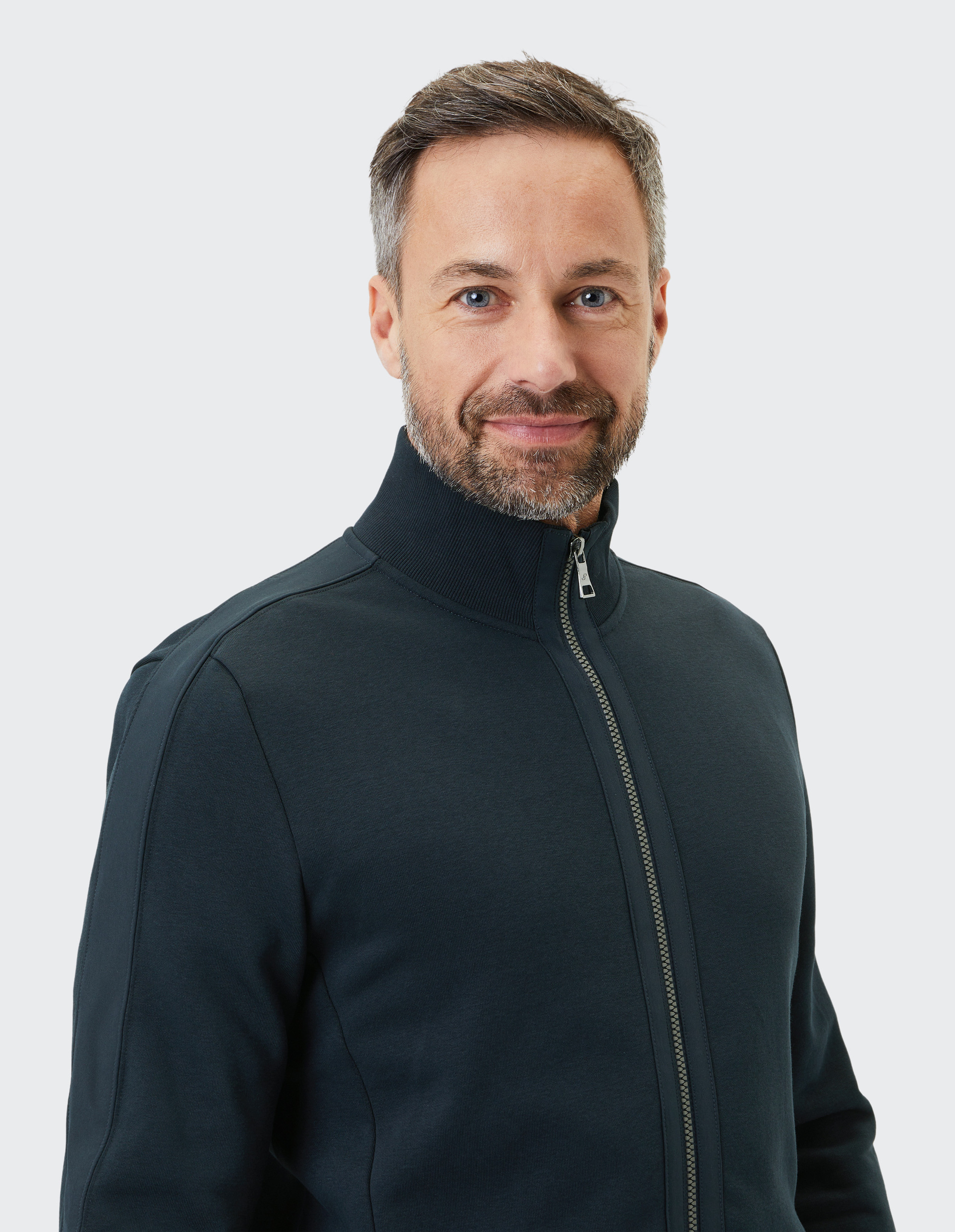 Ein lächelnder Mann mit grauem Haar und Bart trägt eine dunkelblaue Jacke mit Reißverschluss vor einem hellgrauen Hintergrund.