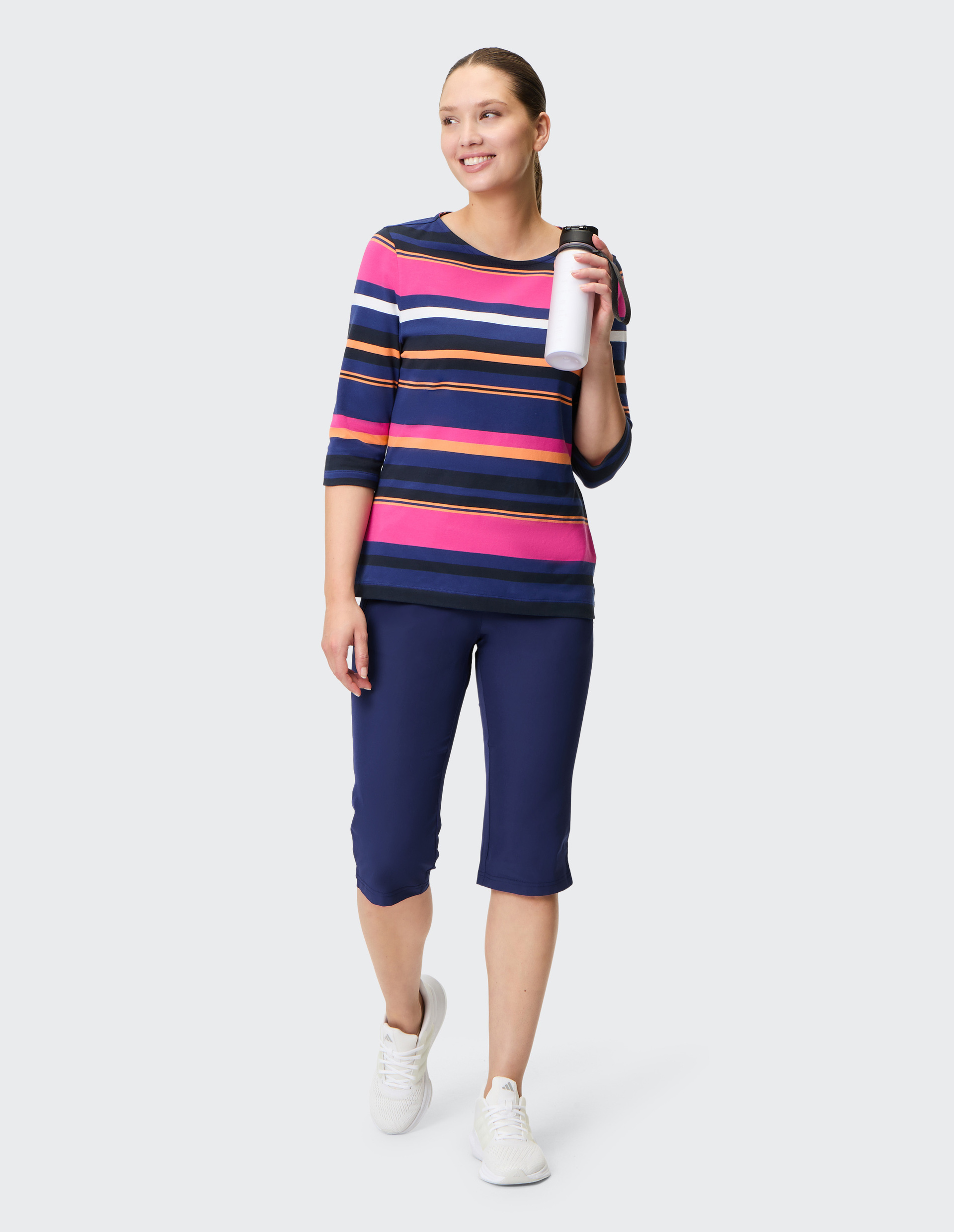 Frau in gestreiftem Shirt und blauen Caprihosen hält eine Trinkflasche, lächelt, steht aufrecht, trägt weiße Turnschuhe.