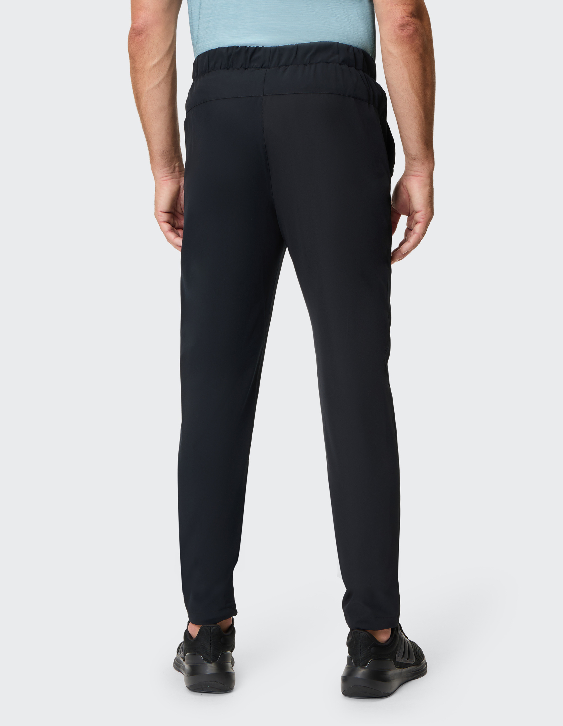 Rückansicht eines Mannes in schwarzer Jogginghose und grauem T-Shirt, der schwarze Sneaker trägt, vor neutralem Hintergrund.