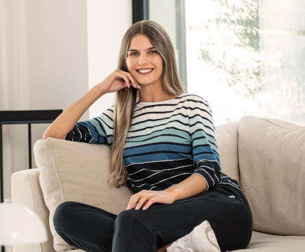 Auf dem Sofa sitzende Frau in dunkelblauer Hose und weiß, hellblau, blau und dunkelblau gestreiftem 3/4-Arm-Shirt der Marke JOY sportswear.
