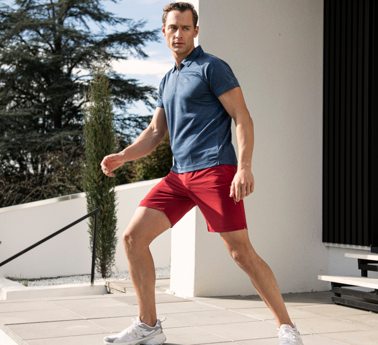 Mann in dunkelblau-meliertem Zip-Poloshirt und roter Shorts der Marke JOY sportswear in sportlicher Pose.