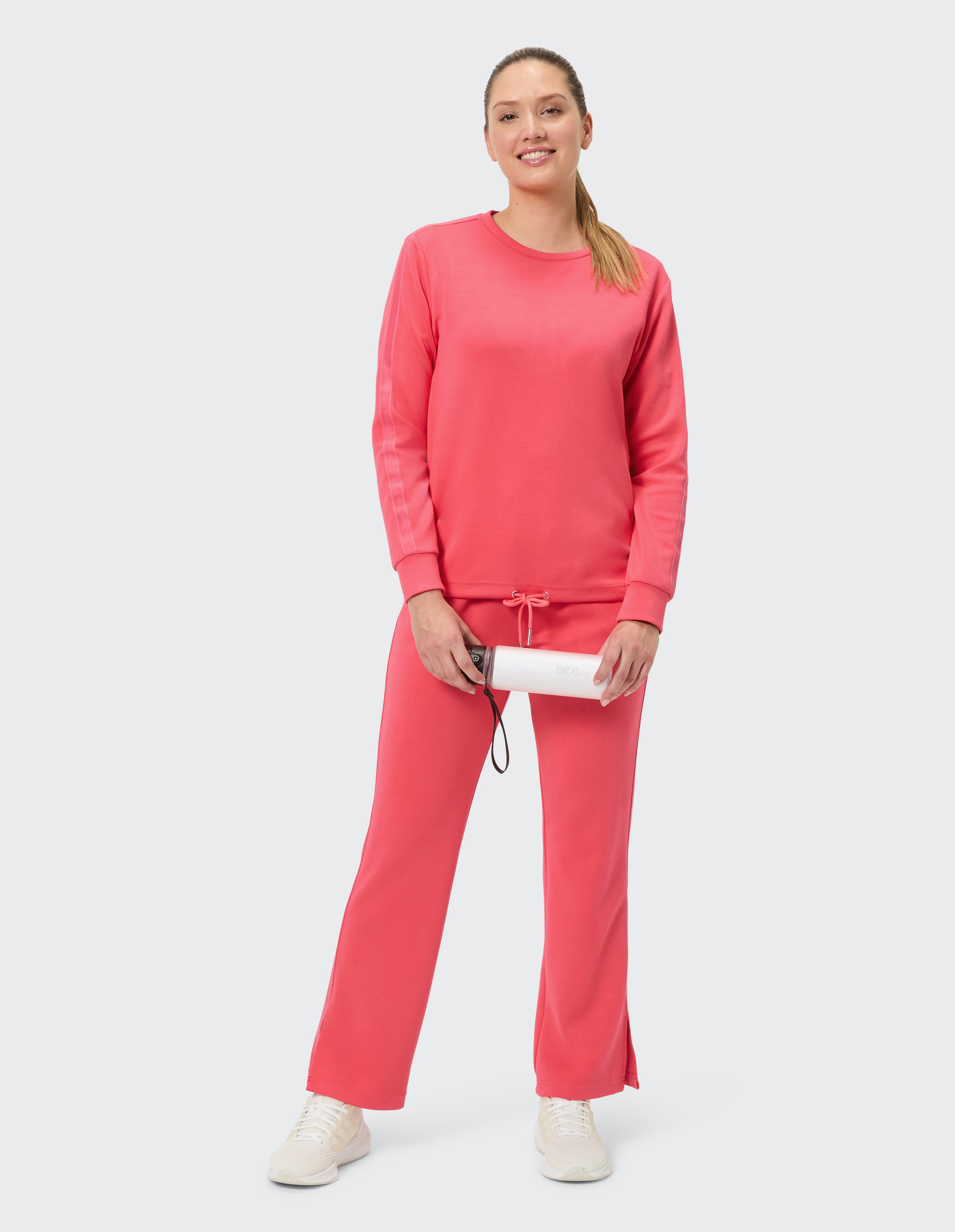 Frau in einem gemütlichen, korallroten Trainingsanzug mit langen Ärmeln und weitem Bein, trägt weiße Sneaker, lächelt leicht.