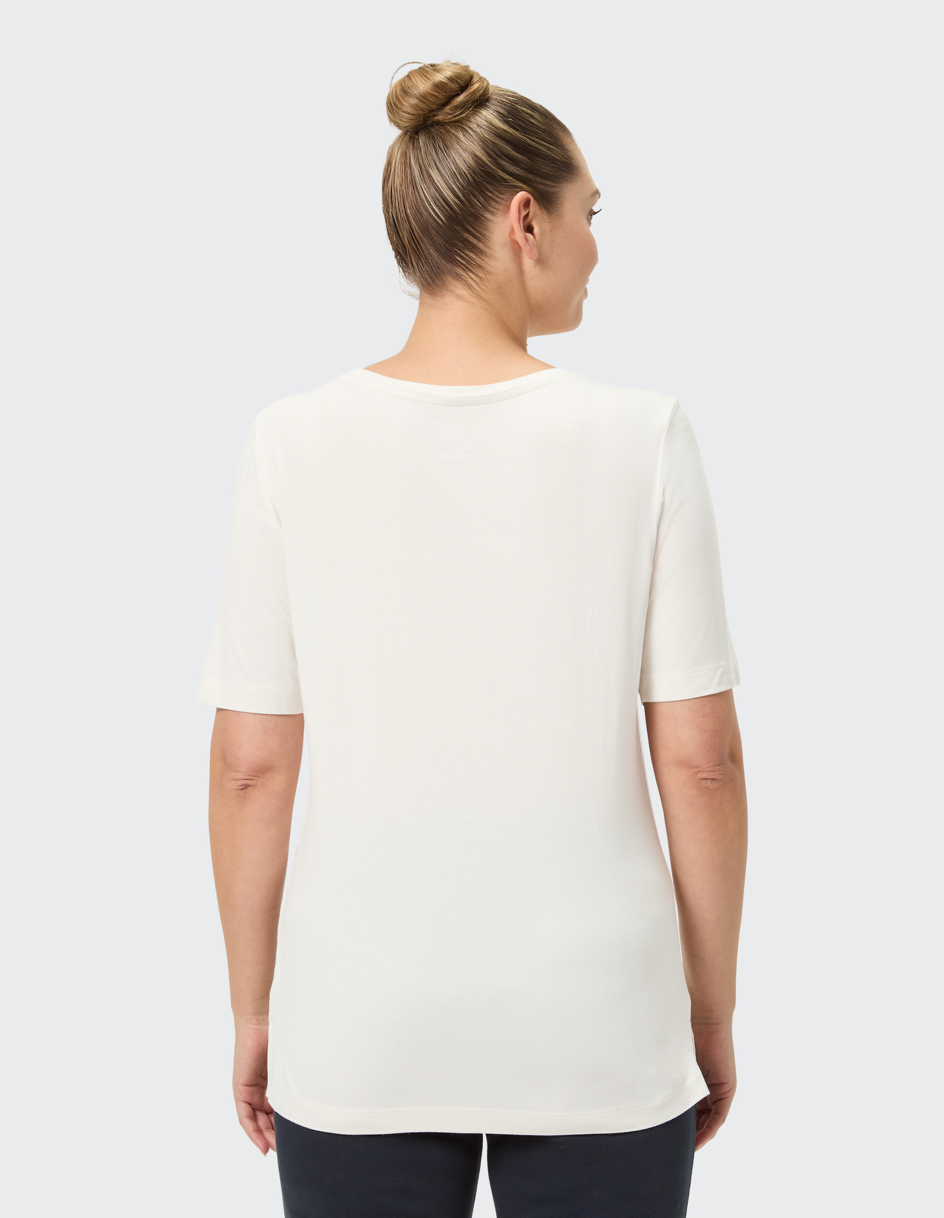 Frau mit Haaren im Dutt trägt ein weißes T-Shirt und steht mit dem Rücken zur Kamera vor grauem Hintergrund.