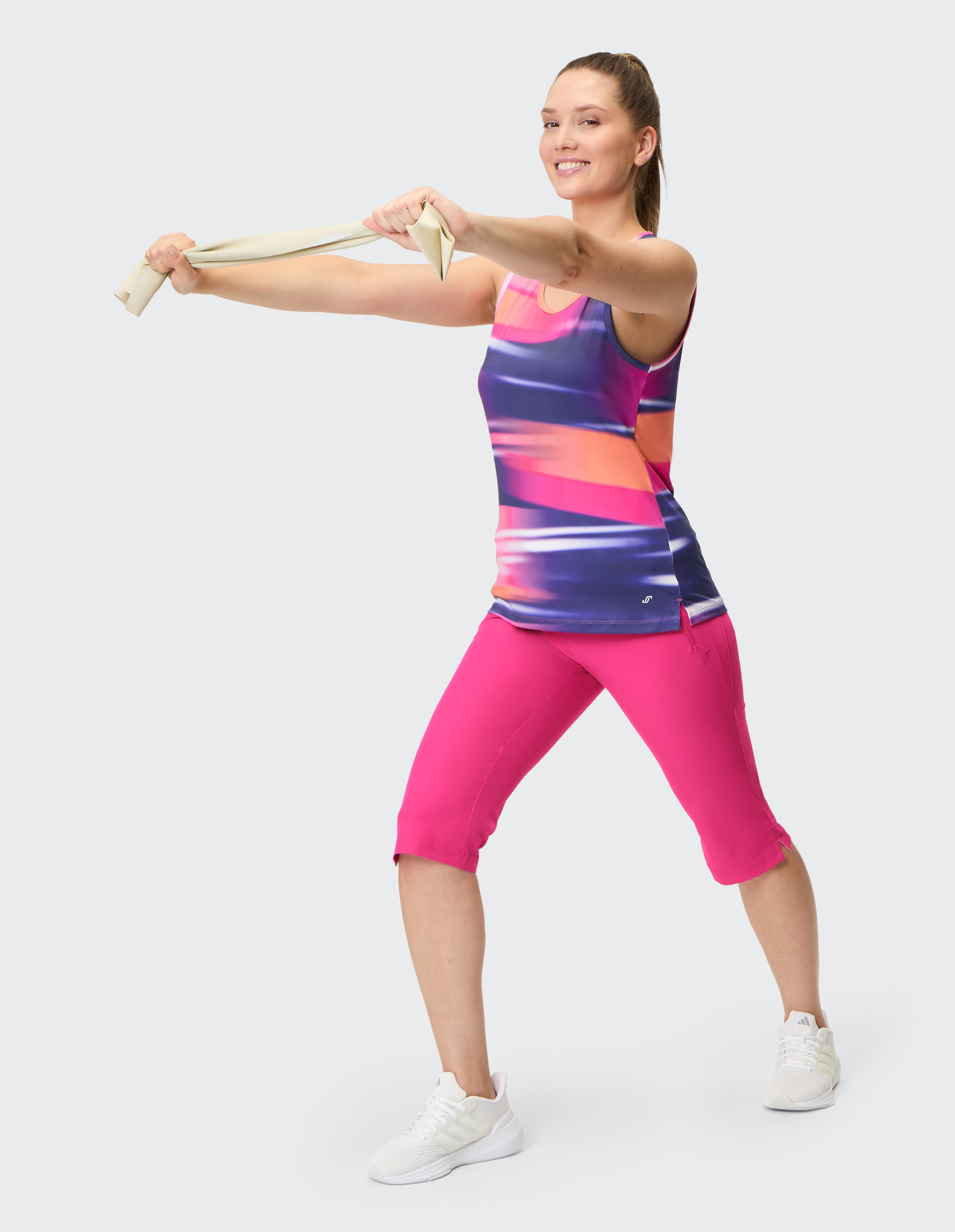 Frau im bunten Sportoutfit streckt mit einem Widerstandsband in Händen die Arme nach vorne, positioniert in Schrittstellung.