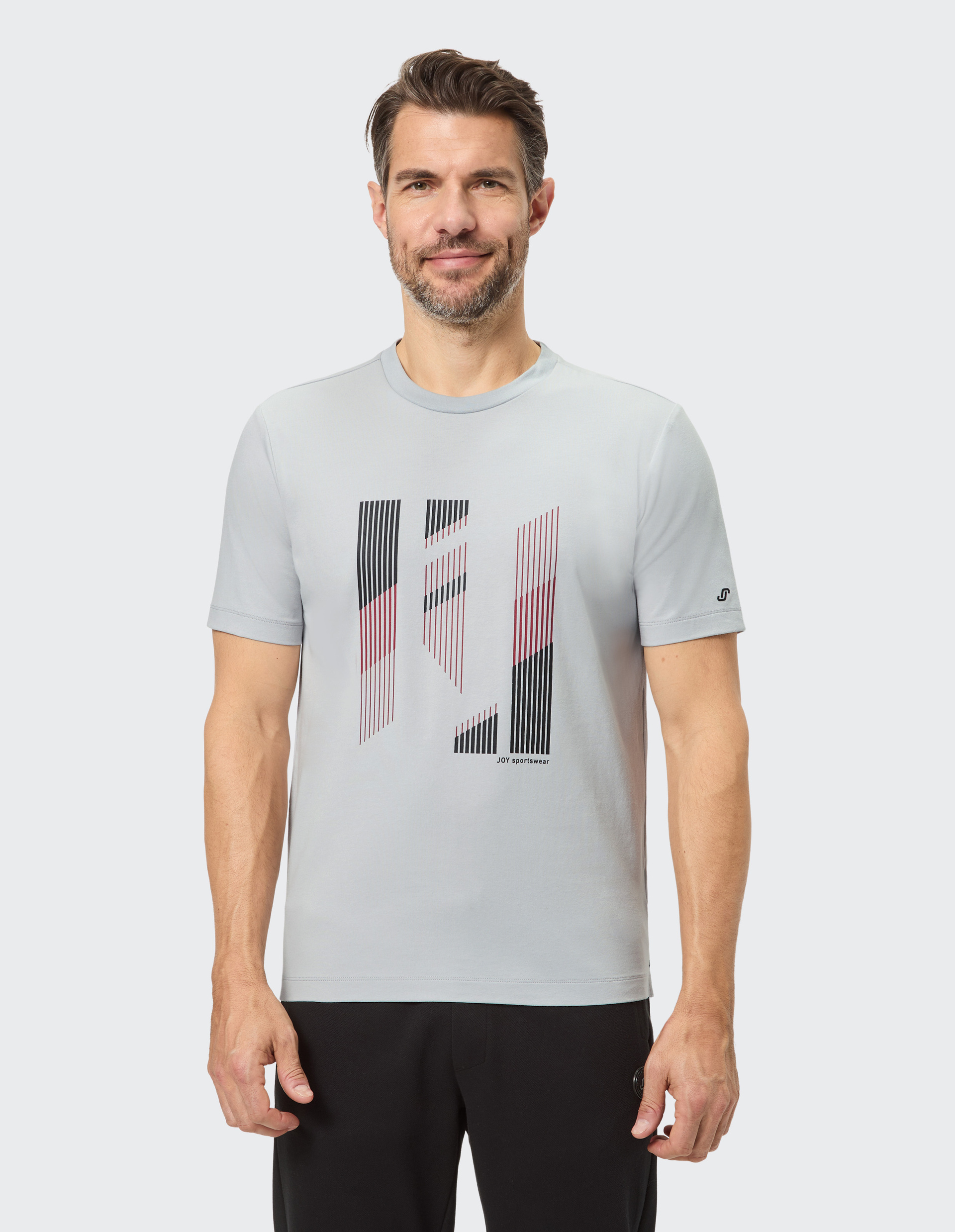 Mann in grauem T-Shirt mit abstraktem Muster in Schwarz und Rot, lächelt leicht, hat kurzen Bart und steht vor neutralem Hintergrund.