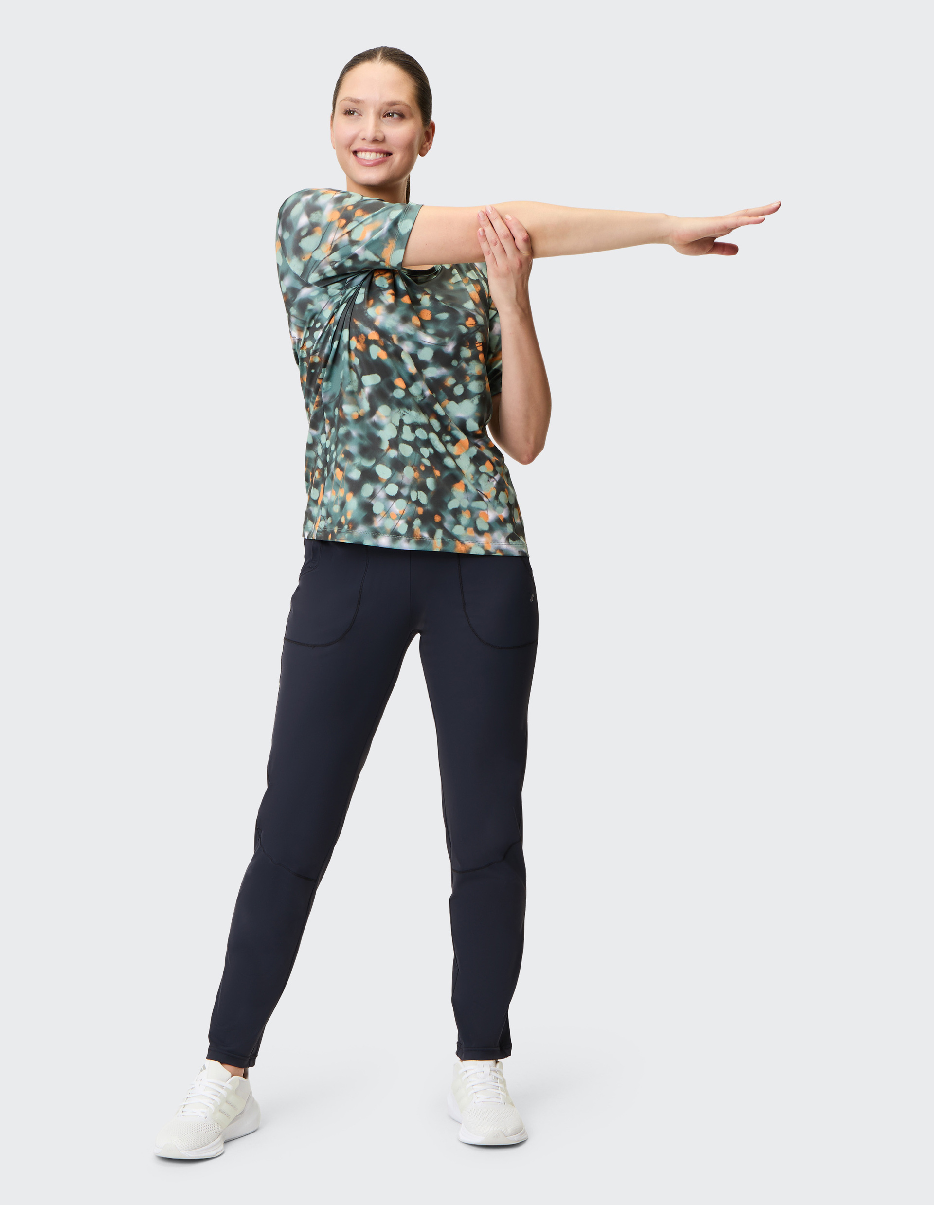 Frau in gemustertem Shirt und dunkler Hose dehnt im Stehen den rechten Arm quer über die Brust, lächelt, auf hellem Hintergrund.