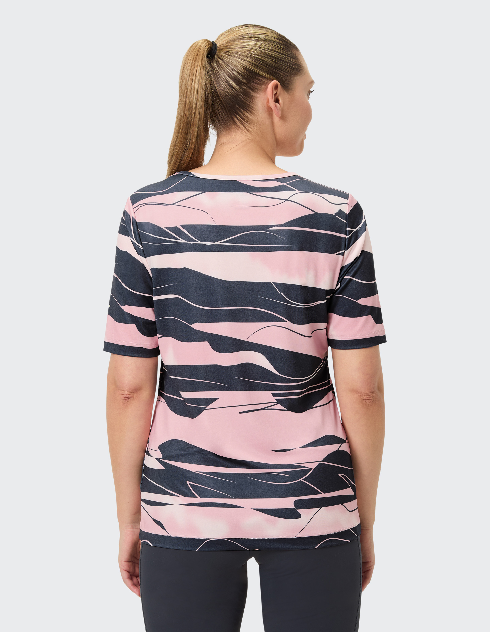 Frau von hinten mit hellem Pferdeschwanz trägt ein rosa-schwarz gemustertes T-Shirt und schwarze Hose vor neutralem Hintergrund.