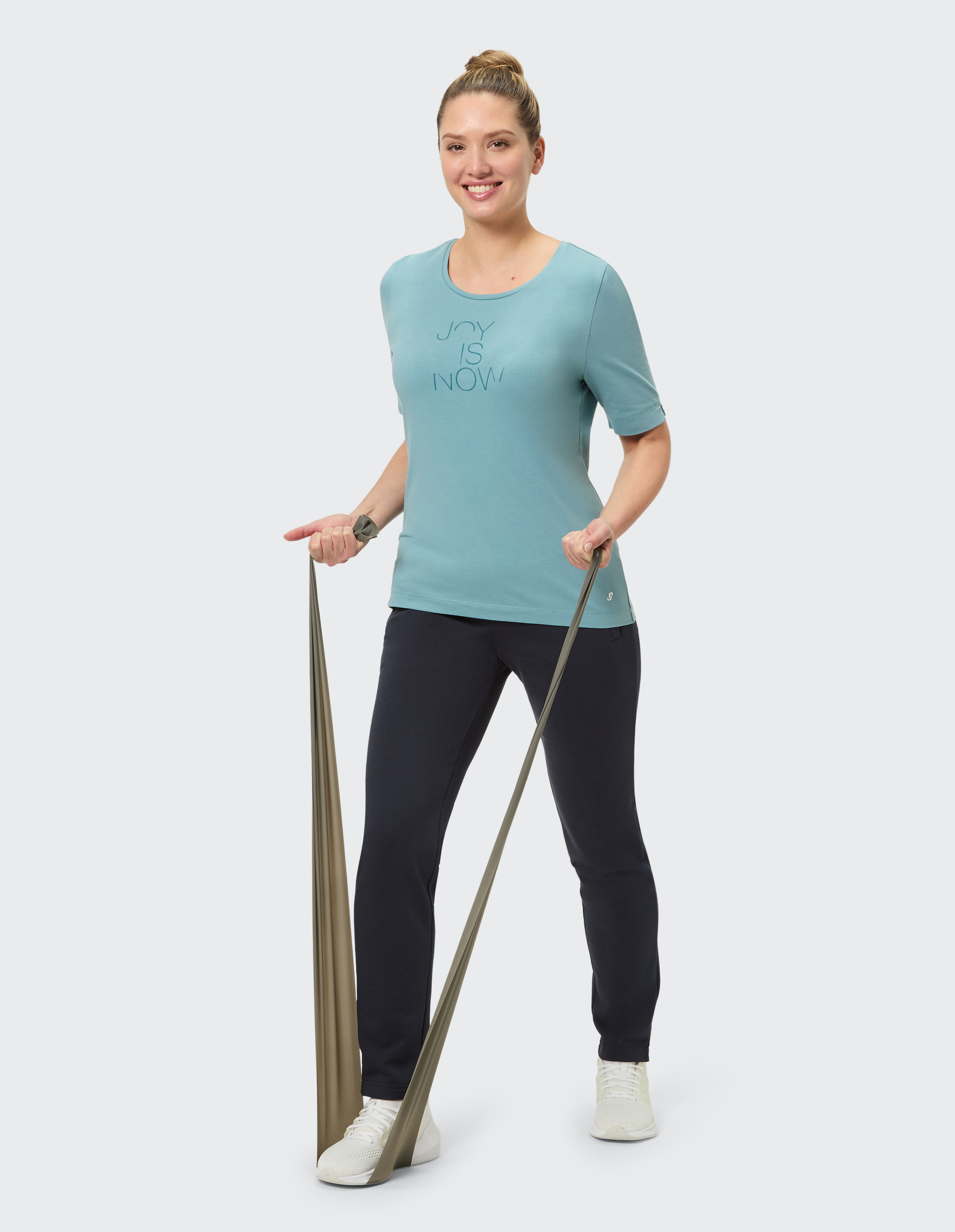 Eine Person trägt ein hellblaues T-Shirt mit der Aufschrift Joy is Now, dunkelblaue Hosen und weiße Sneaker, vor grauem Hintergrund.