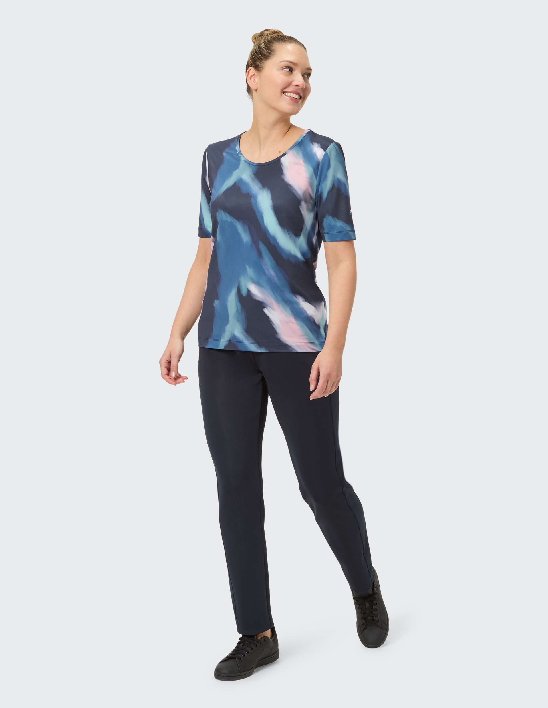 Frau trägt ein bunt gemustertes T-Shirt und schwarze Hose, steht frontal und lächelt leicht vor einem neutralen Hintergrund.