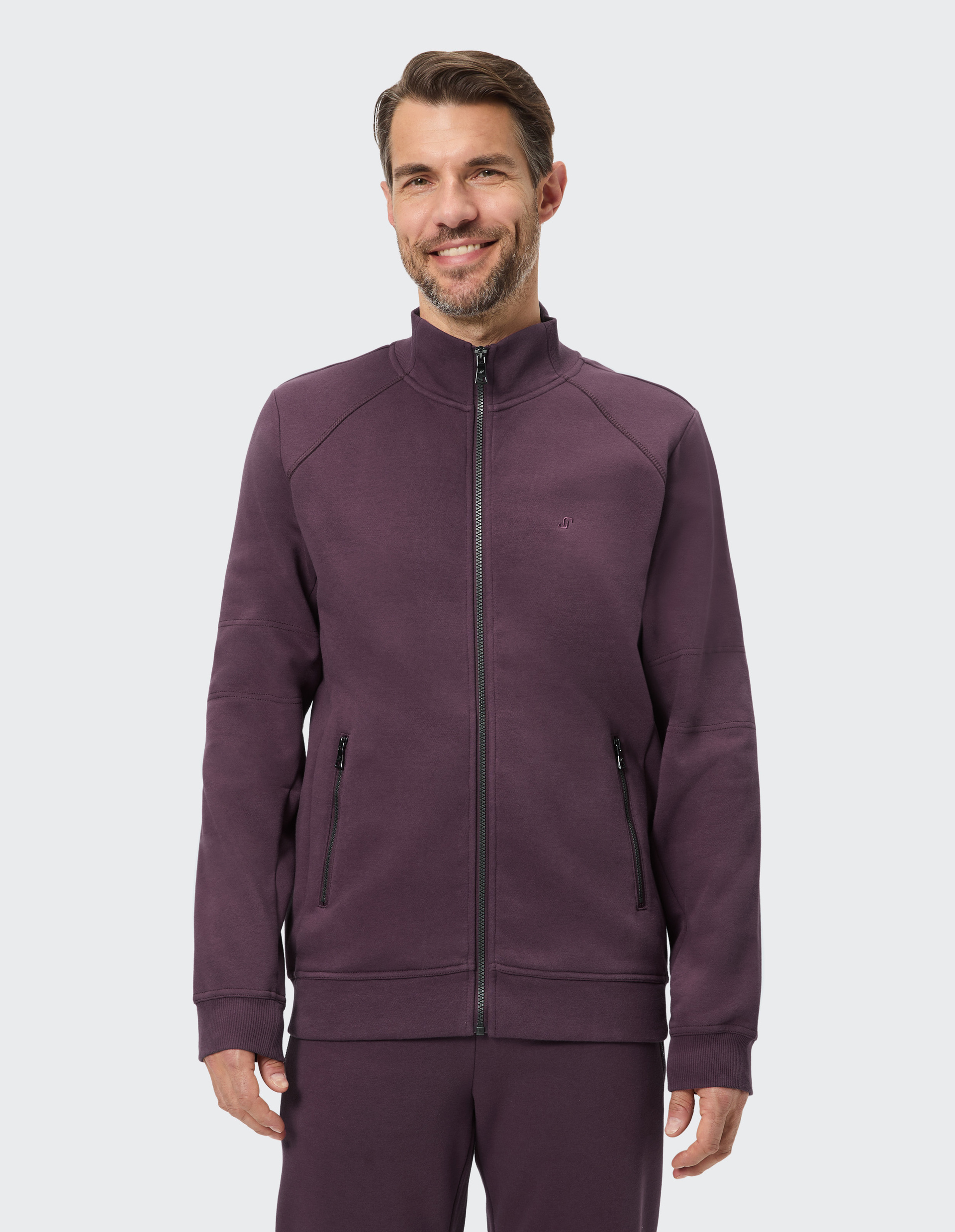 Mann lächelt in die Kamera, trägt eine dunkelviolette Trainingsjacke mit Reißverschluss und Stehkragen vor grauem Hintergrund.