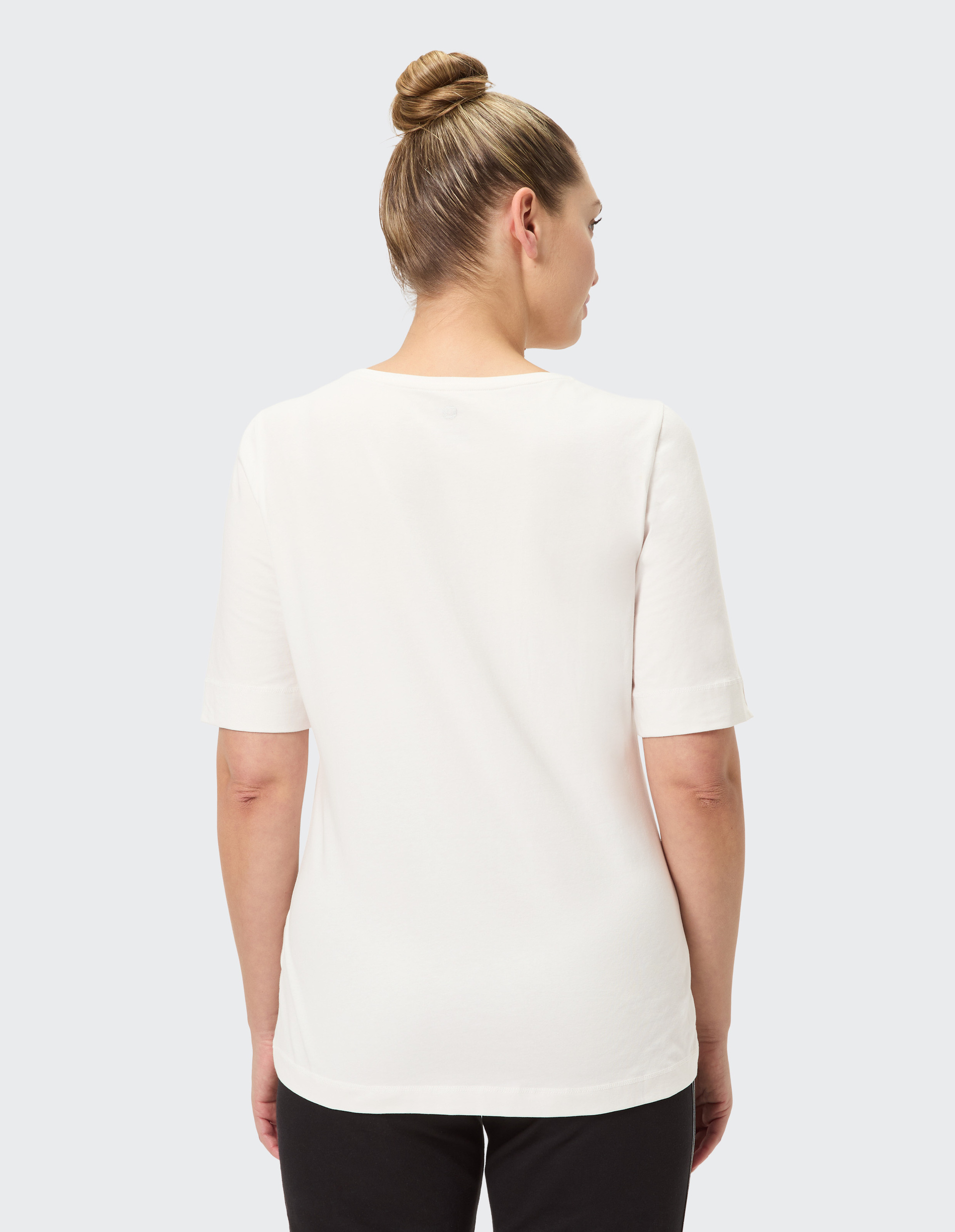 Eine Person steht mit dem Rücken zur Kamera, trägt ein weißes T-Shirt und schwarze Hose, die Haare zu einem Dutt gebunden.