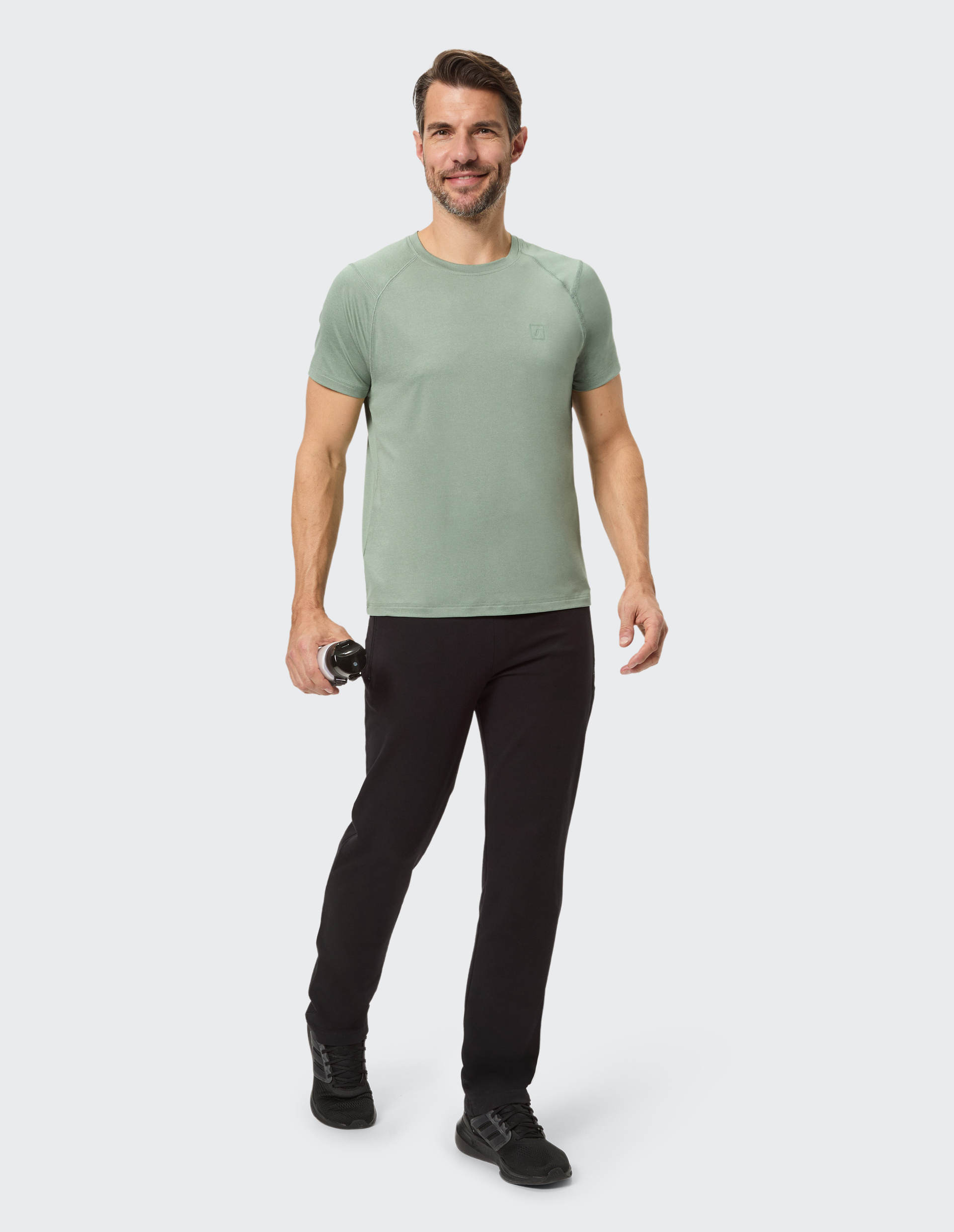 Mann in grünem T-Shirt und schwarzer Hose steht lächelnd vor hellem Hintergrund, beide Hände locker an den Seiten.