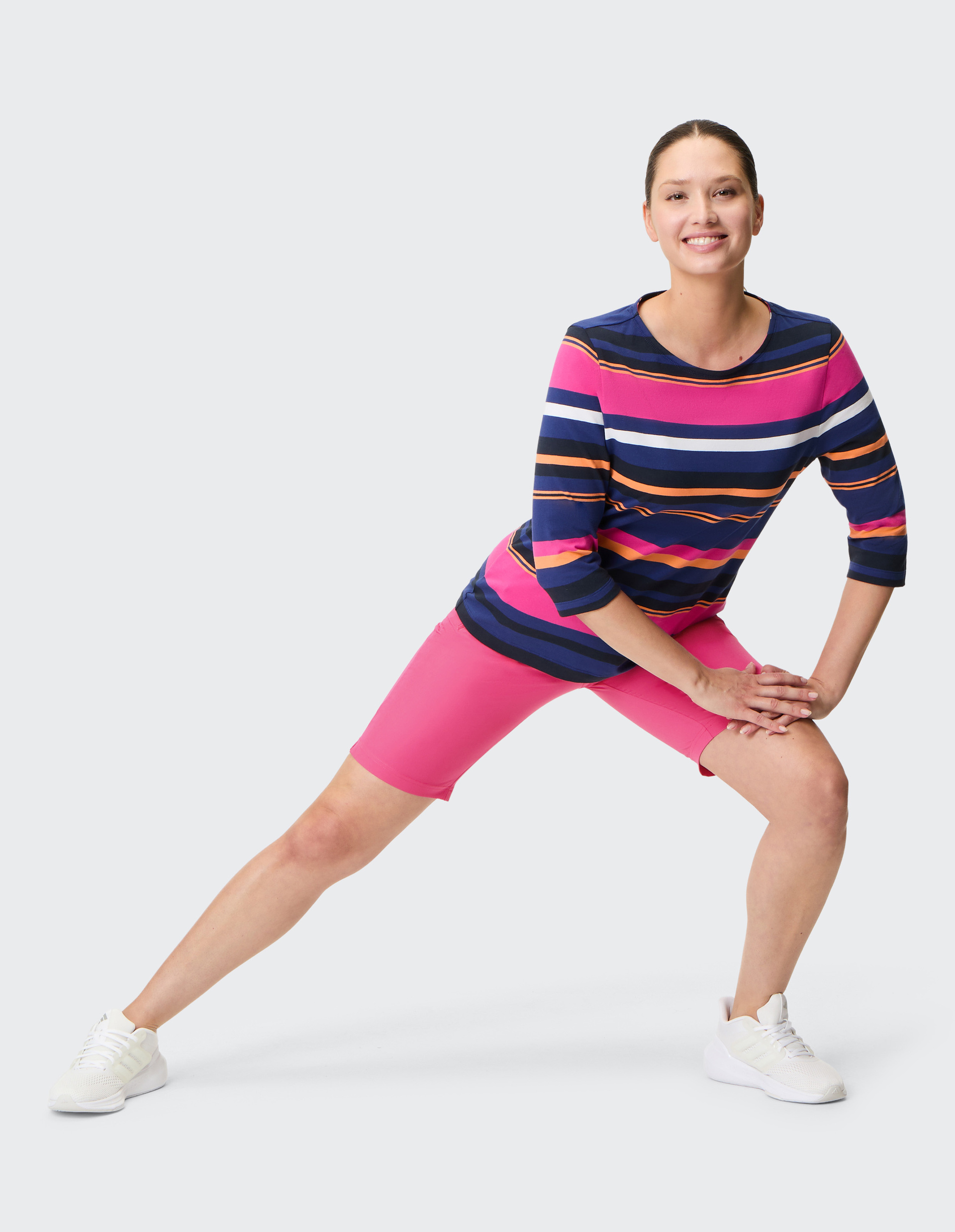 Frau in bunter Streifenbluse und pinker Radlerhose dehnt lächelnd seitwärts das linke Bein, trägt weiße Sneaker.
