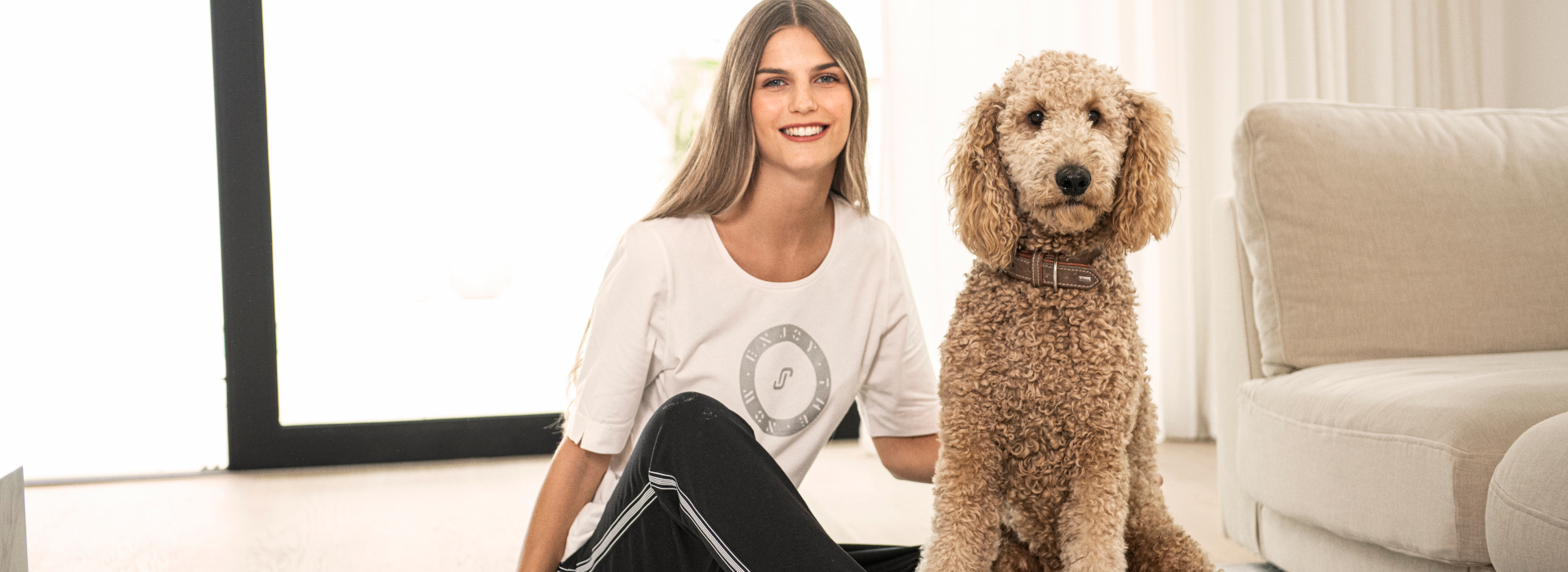 Frau in weißem T-Shirt mit Print der Marke JOY sportswear auf dem Boden sitzend und den Hund neben sich streichelnd.