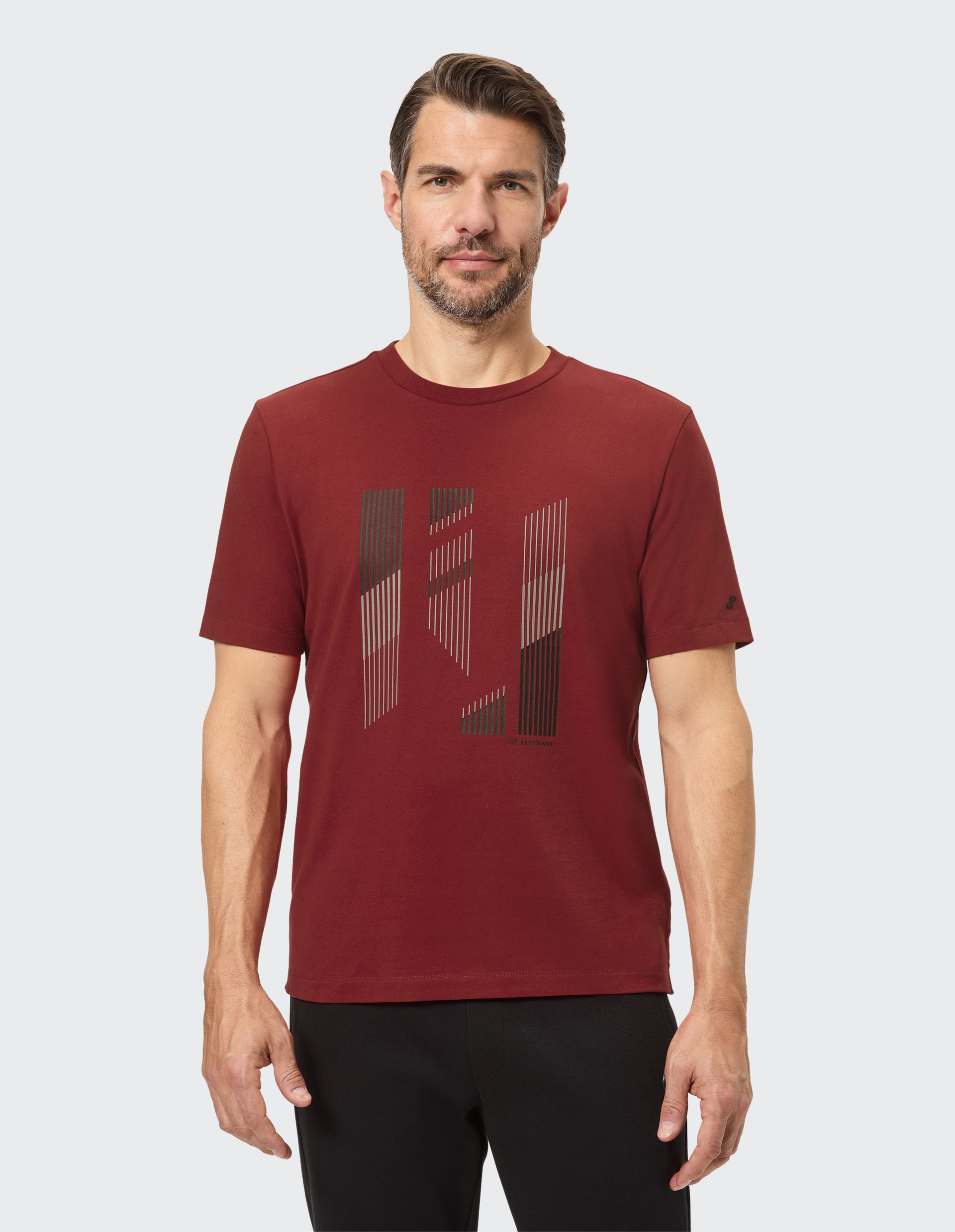 Ein Mann trägt ein dunkelrotes T-Shirt mit einem grafischen, vertikal gestreiften Muster und schwarzen Hosen vor weißem Hintergrund.