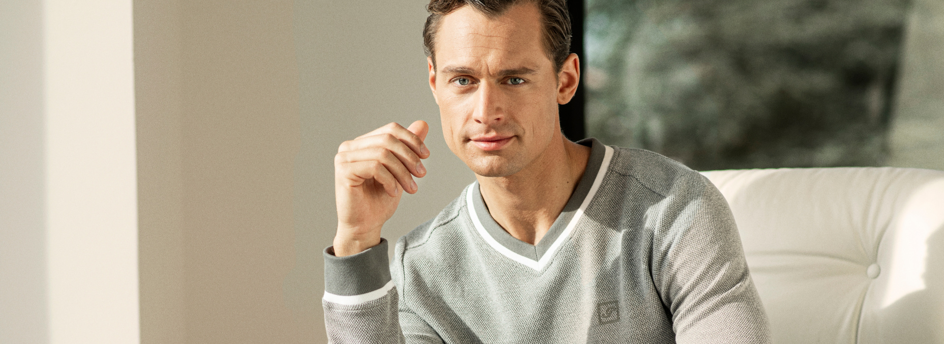 Mann mit grauem Sweatshirt der Marke JOY sportswear sitzt auf einem weißen Sessel.