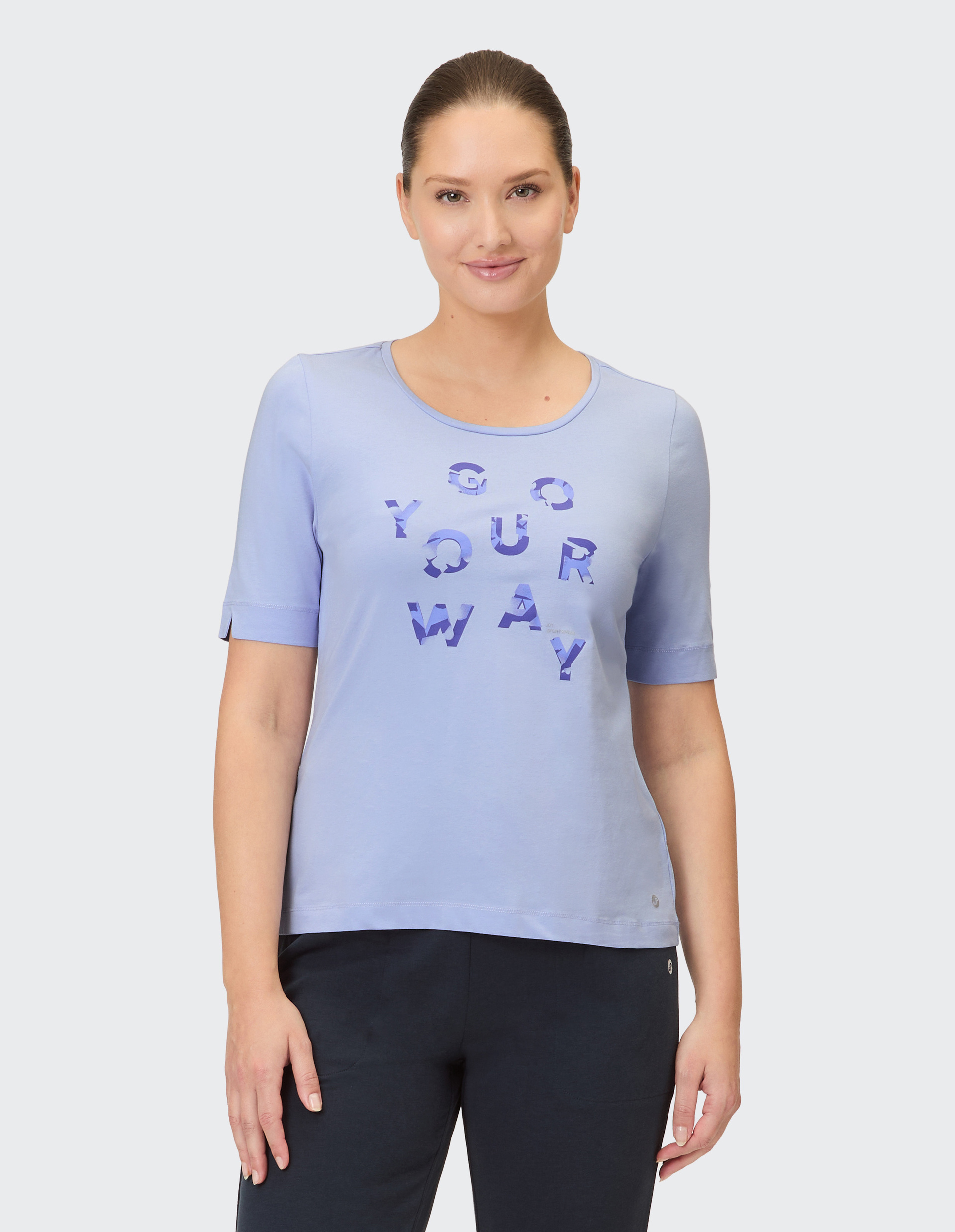 Eine Frau trägt ein hellblaues T-Shirt mit der Aufschrift "GO YOUR WAY" in lila, die Form ist kurzärmelig und rundhalsausschnitt.