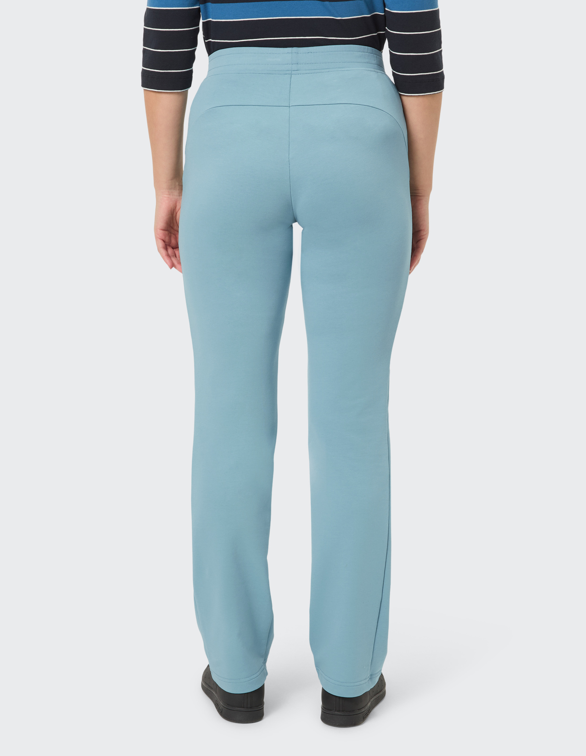 Person in hellblauer Hose und gestreiftem Oberteil, von hinten gesehen, trägt schwarze Schuhe auf hellem Hintergrund.