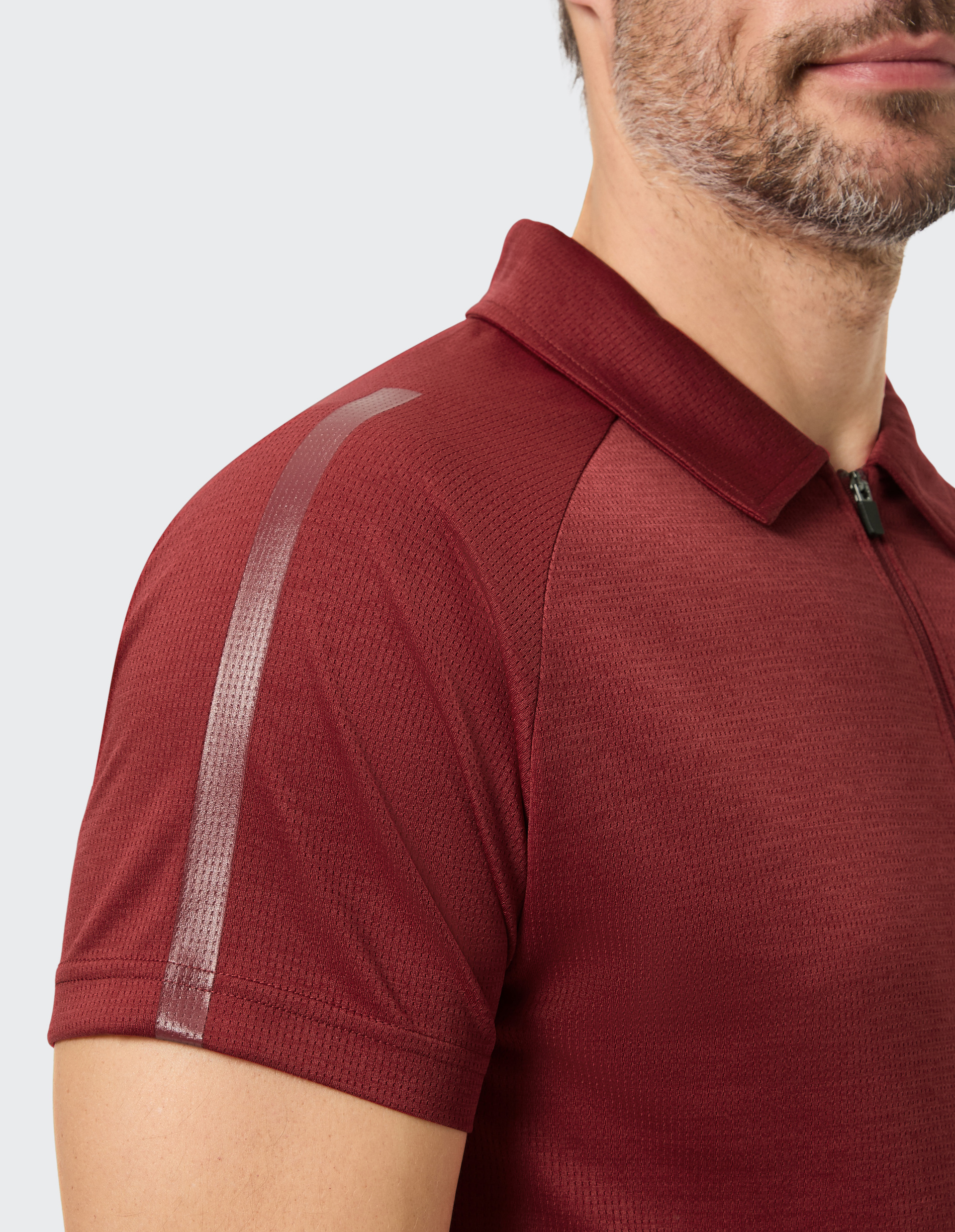 Ein Mann trägt ein weinrotes Poloshirt mit Reißverschluss und reflektierenden Streifen auf der Schulter, Bart im Anschnitt sichtbar.