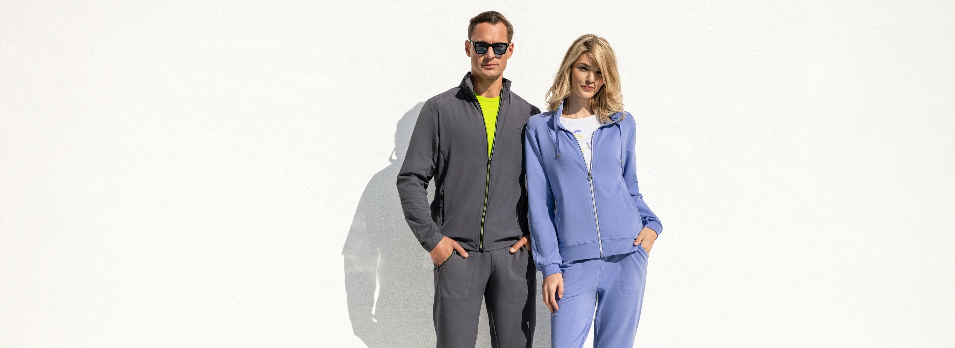 Frau in blauem Anzug steht neben Mann in grauem Anzug der Marke JOY sportswear. Vor den beiden ist die Ecke eines Pools zu sehen.