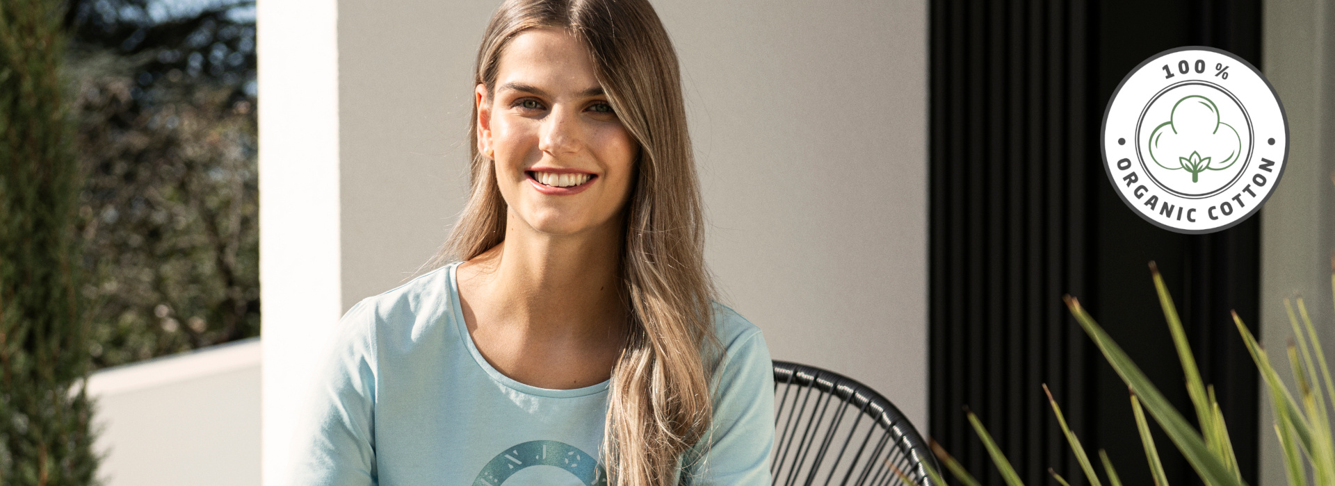 Frau in hellblauem T-Shirt der Marke JOY sportswear sieht lächelnd nach vorne. In der Ecke des Bildes ist das Zeichen für Organic Cotton angebracht.