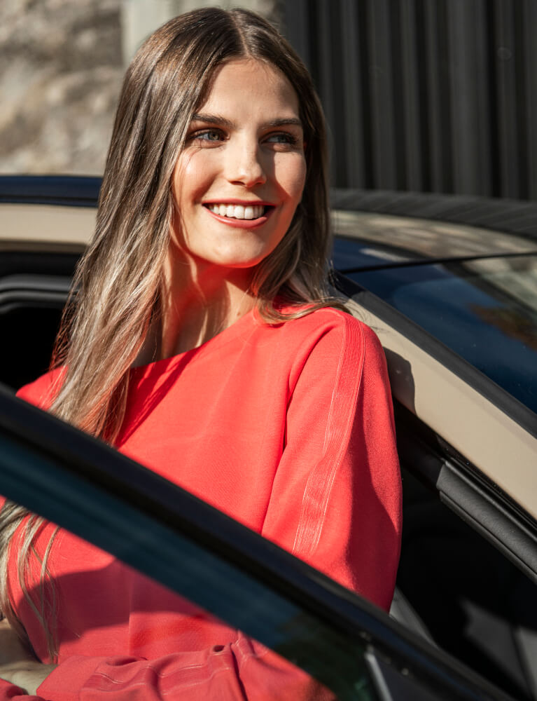 Frau in rotem Sweatshirt der Marke JOY sportswear lehnt an beigenem Auto.