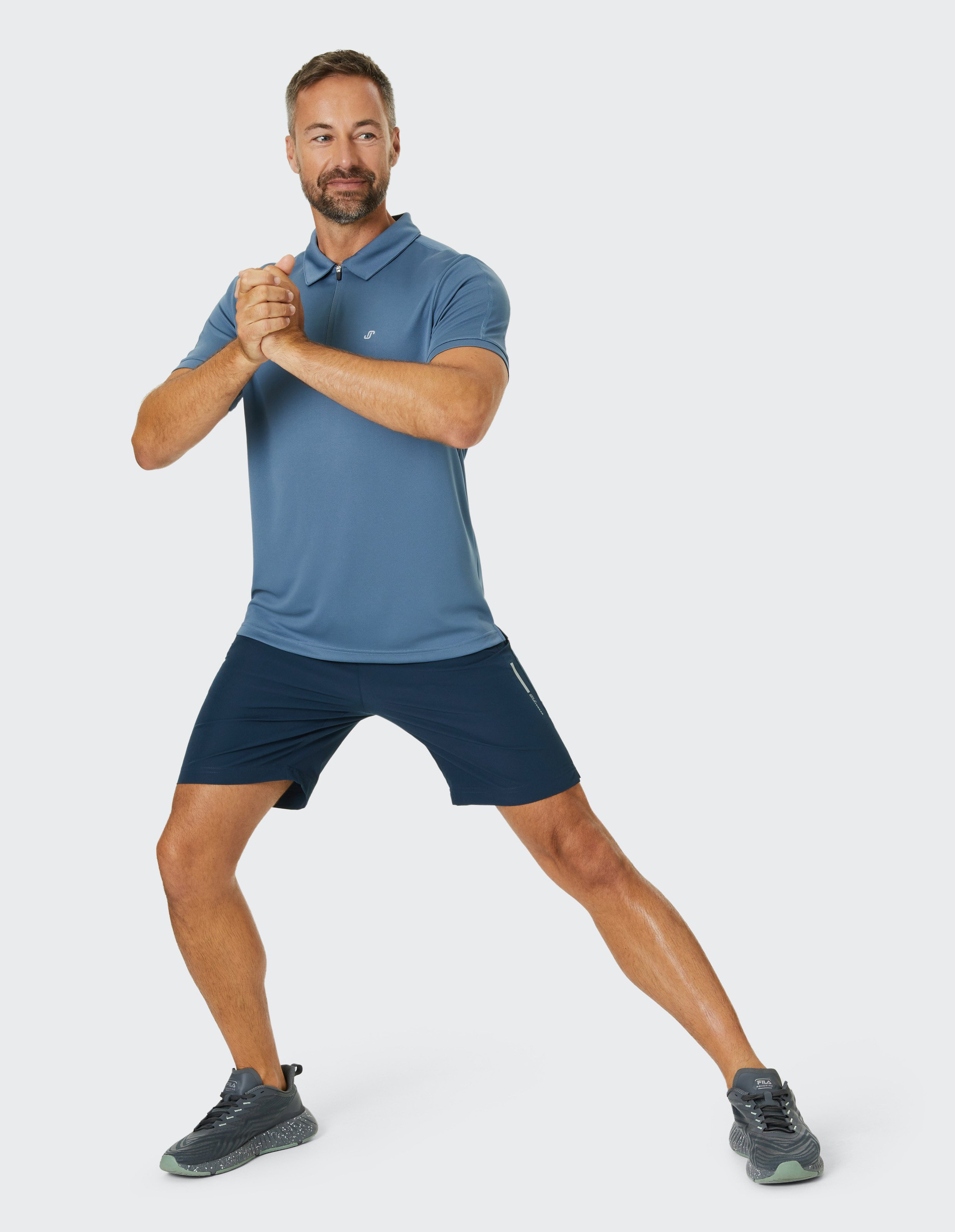 Ein Mann in blauer Sportkleidung macht einen Ausfallschritt, verschränkt die Hände und trägt graue Sportschuhe.