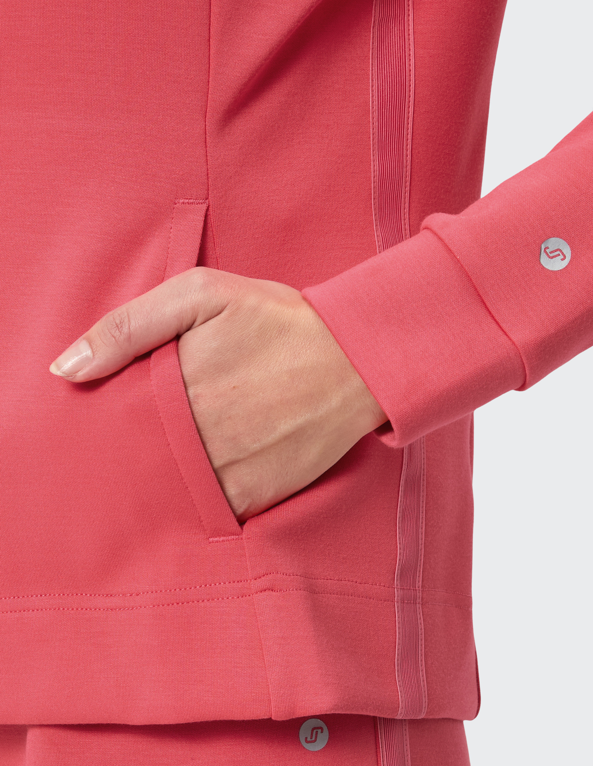 Eine Hand steckt in einer flauschigen, korallroten Jackentasche mit gerippten Details und einem kleinen, kreisförmigen Logo am Ärmel.