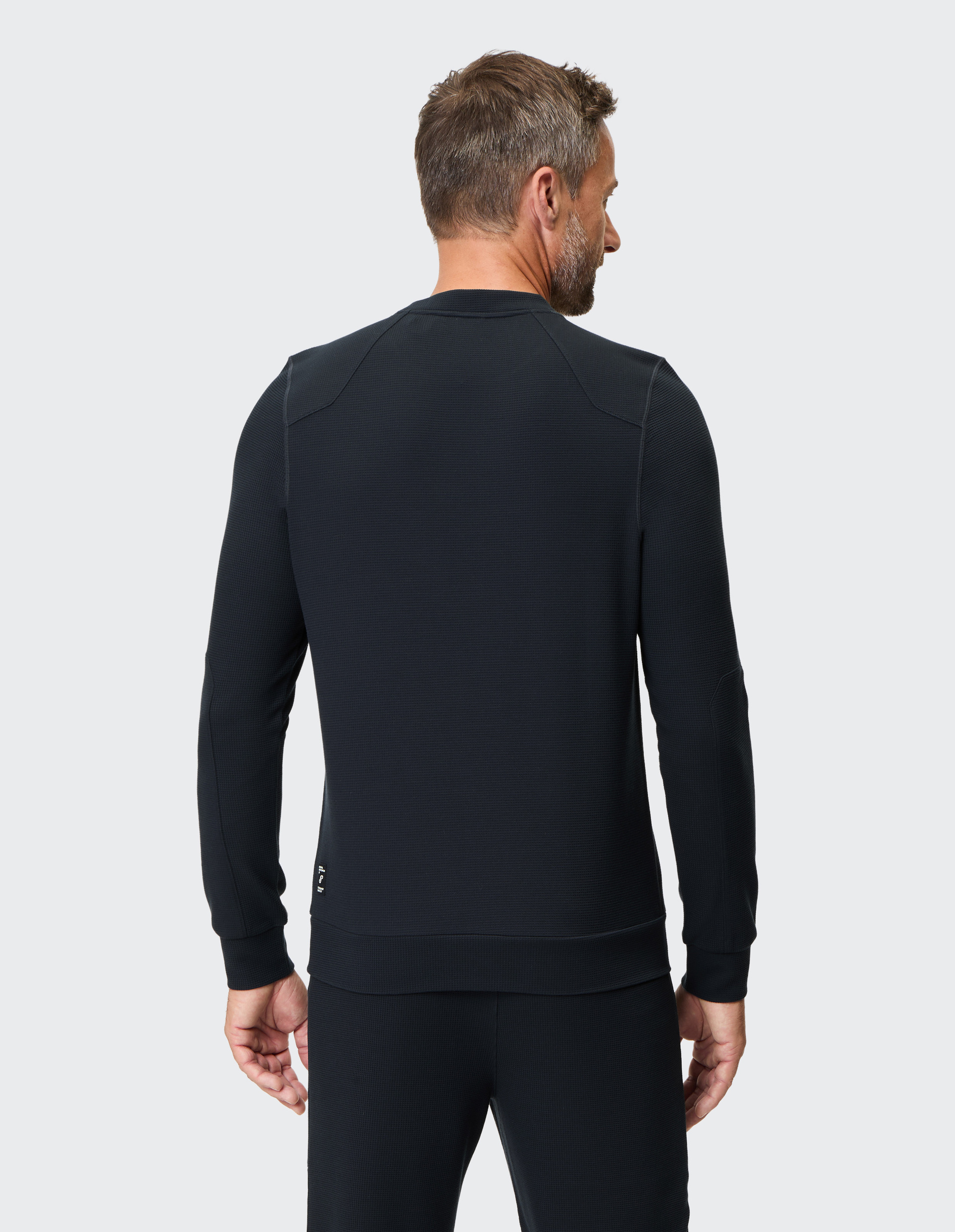 Ein Mann in schwarzem Pullover und Hose steht mit dem Rücken zur Kamera vor einem hellen Hintergrund.