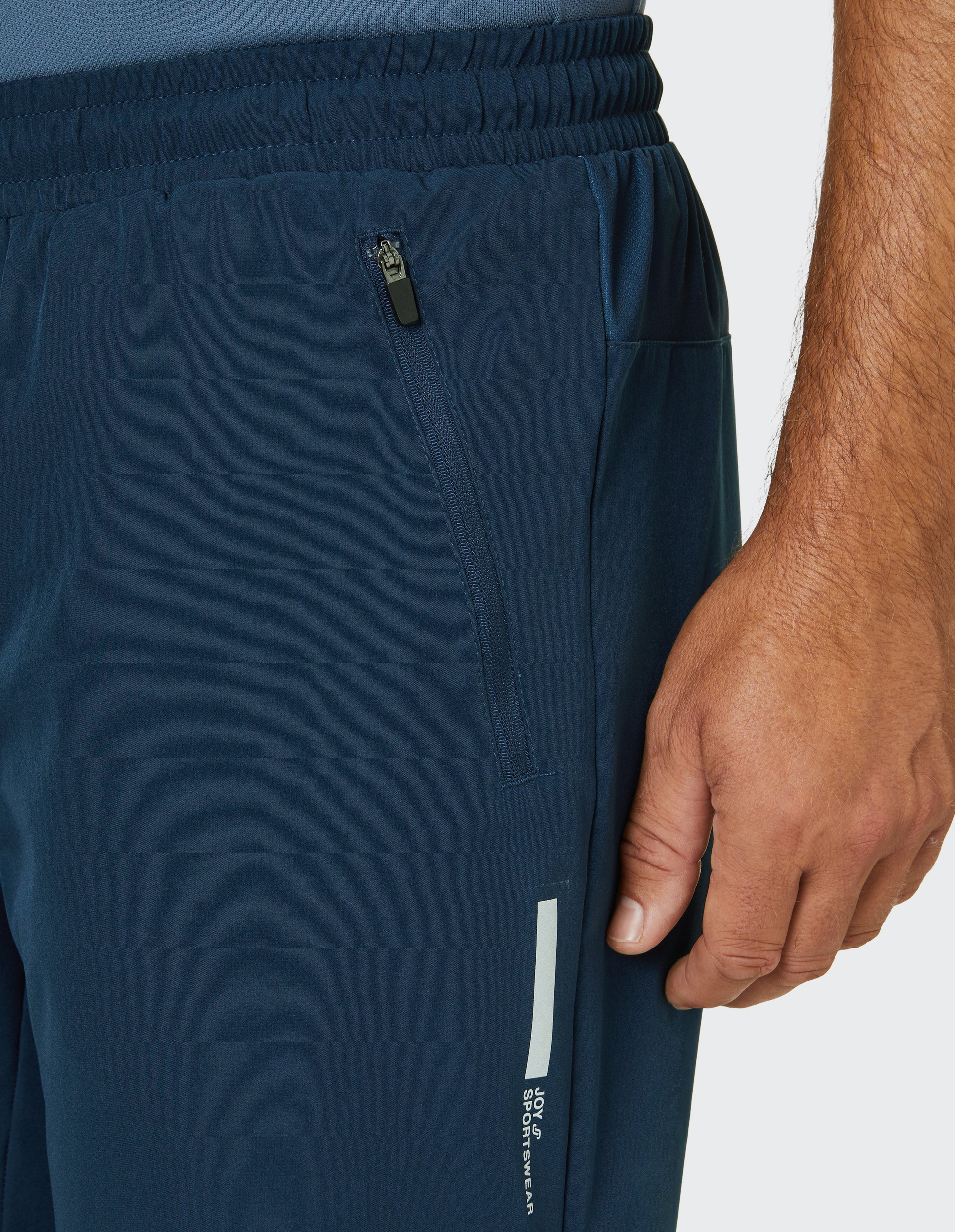 Nahaufnahme einer dunkelblauen Sporthose mit elastischem Bund, Reißverschlusstasche und dezentem Logoaufdruck; Hand eines Mannes daneben.