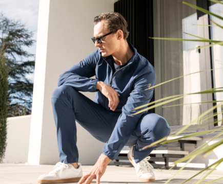 In der Hocke sitzender Mann mit Sonnenbrille in blauem Jogginganzug der Marke JOY sportswear.