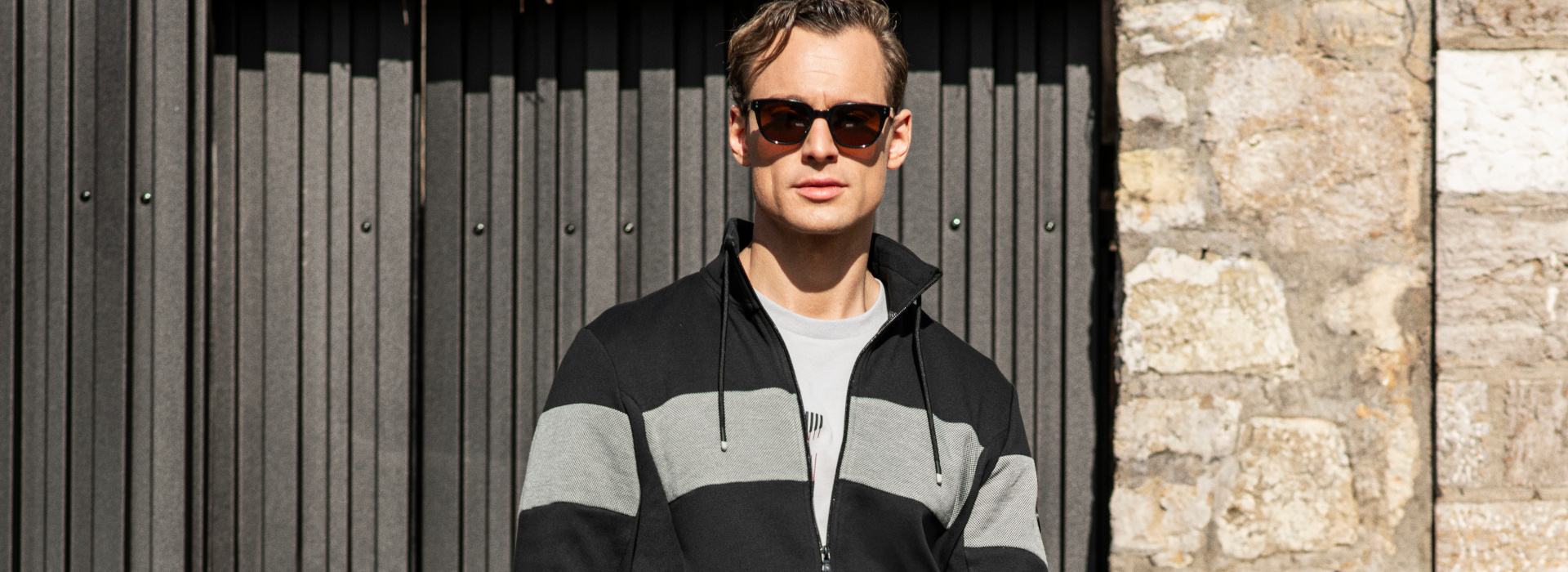 Mann steht vor einer Garage und trägt eine Sonnenbrille und eine gestreifte Jacke der Marke JOY sportswear.