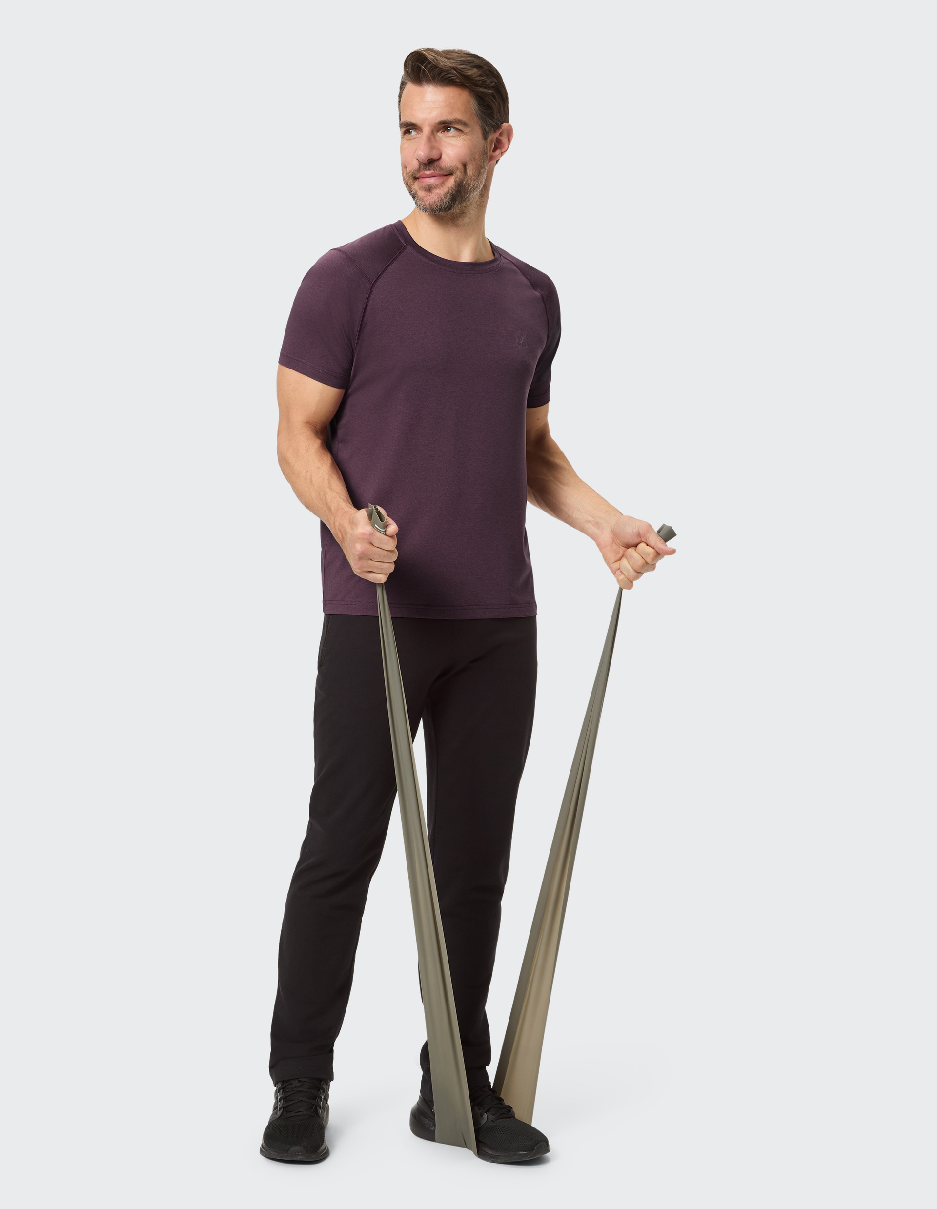 Ein Mann in einem lila T-Shirt und schwarzen Hosen steht vor neutralem Hintergrund, lächelt leicht und schaut direkt in die Kamera.