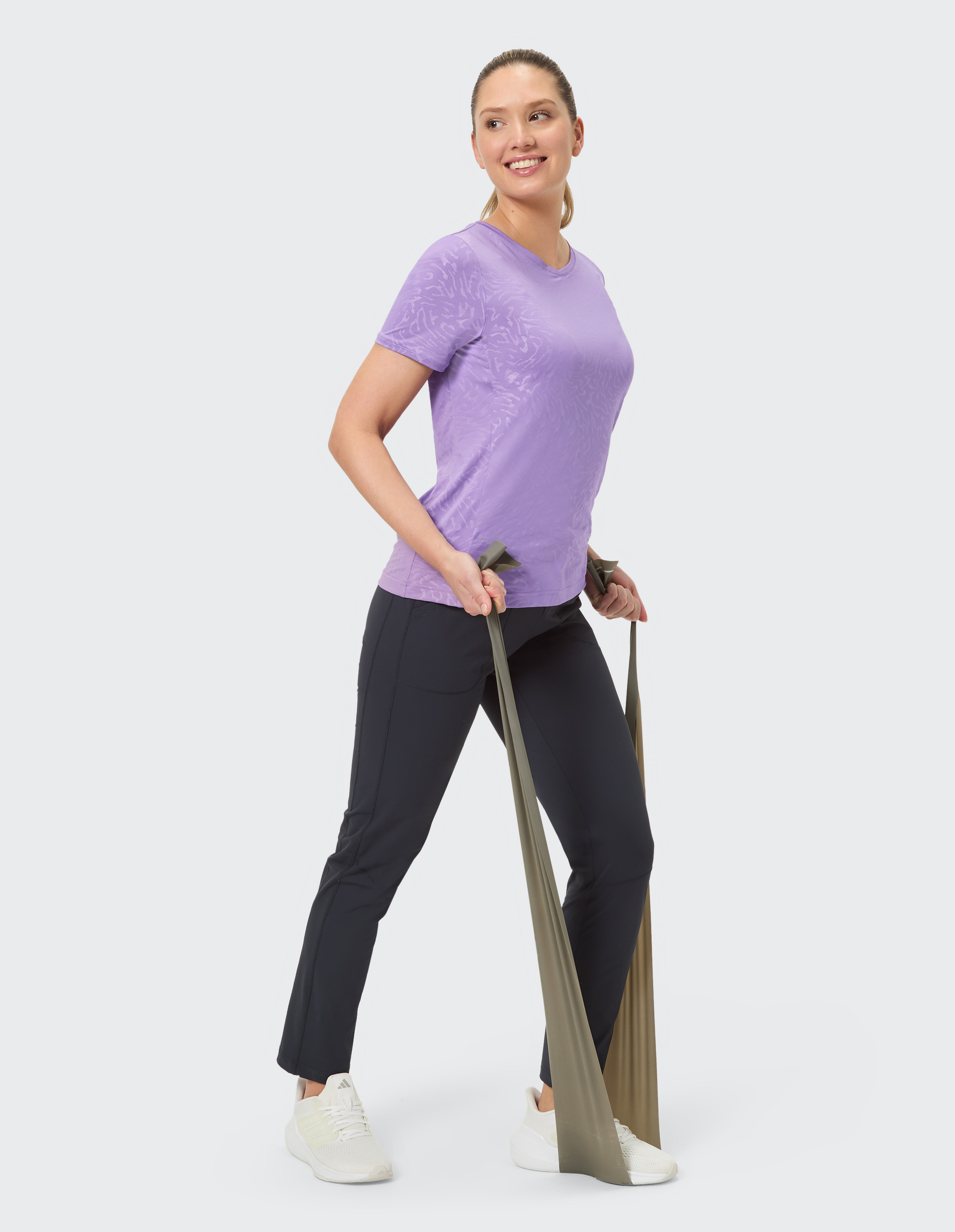 Frau in lila T-Shirt und schwarzen Hosen steht frontal, lächelnd, mit weißem Hintergrund. Ihre Haare sind zurückgebunden.
