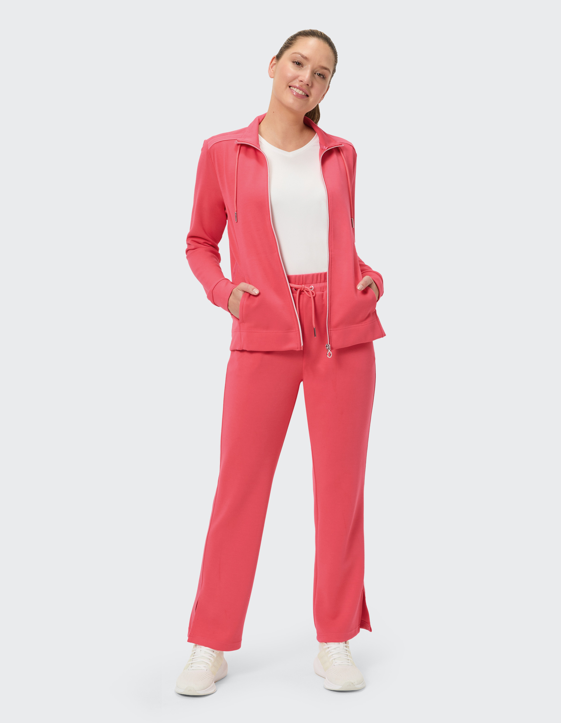 Person in einem pinken Jogginganzug mit Reißverschlussjacke und Trainingshose, trägt weiße Turnschuhe, steht frontal vor einem weißen Hintergrund.