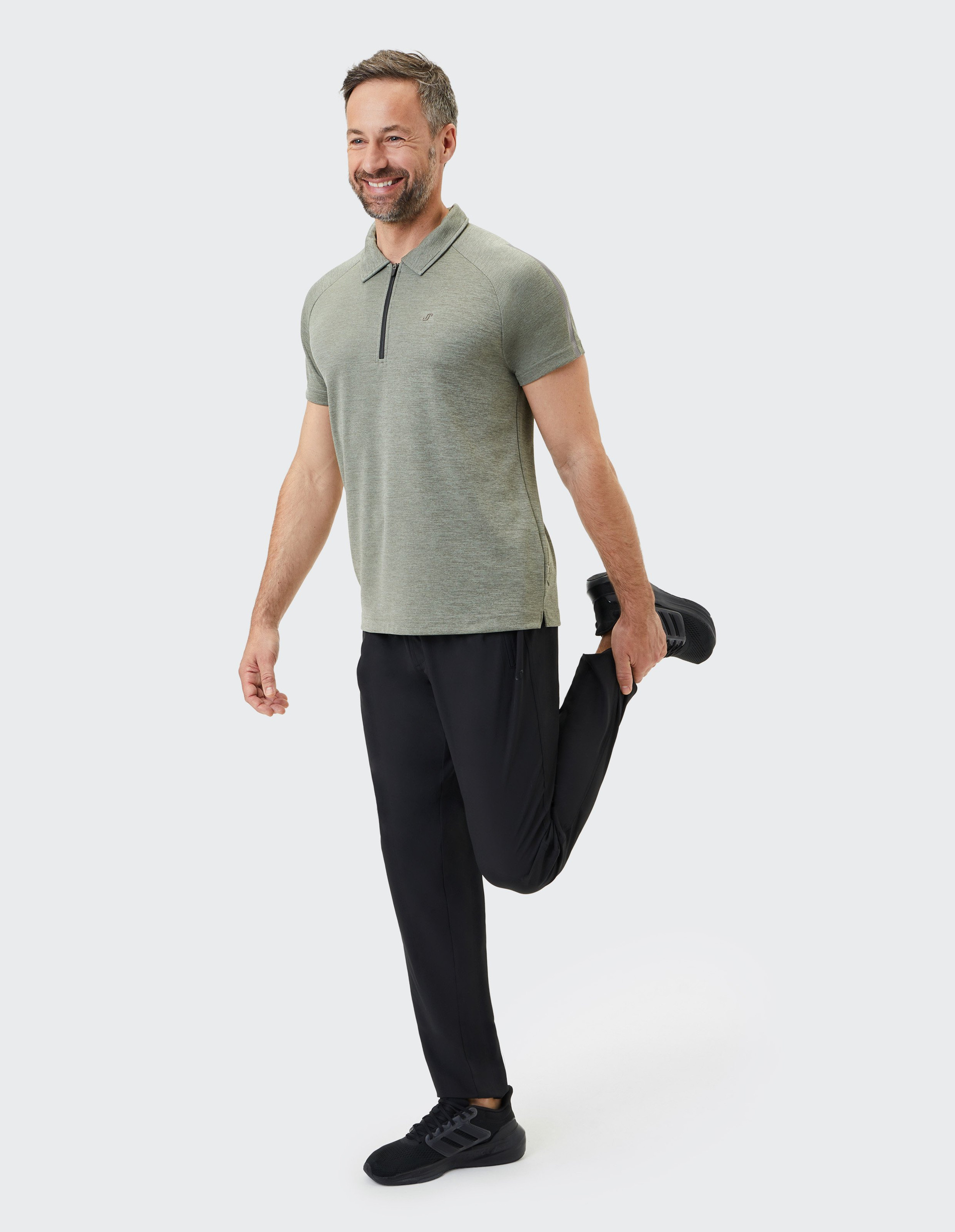 Mann in grauem Poloshirt und schwarzer Hose, lächelnd, dehnt eine Beinmuskulatur stehend, auf weißem Hintergrund.