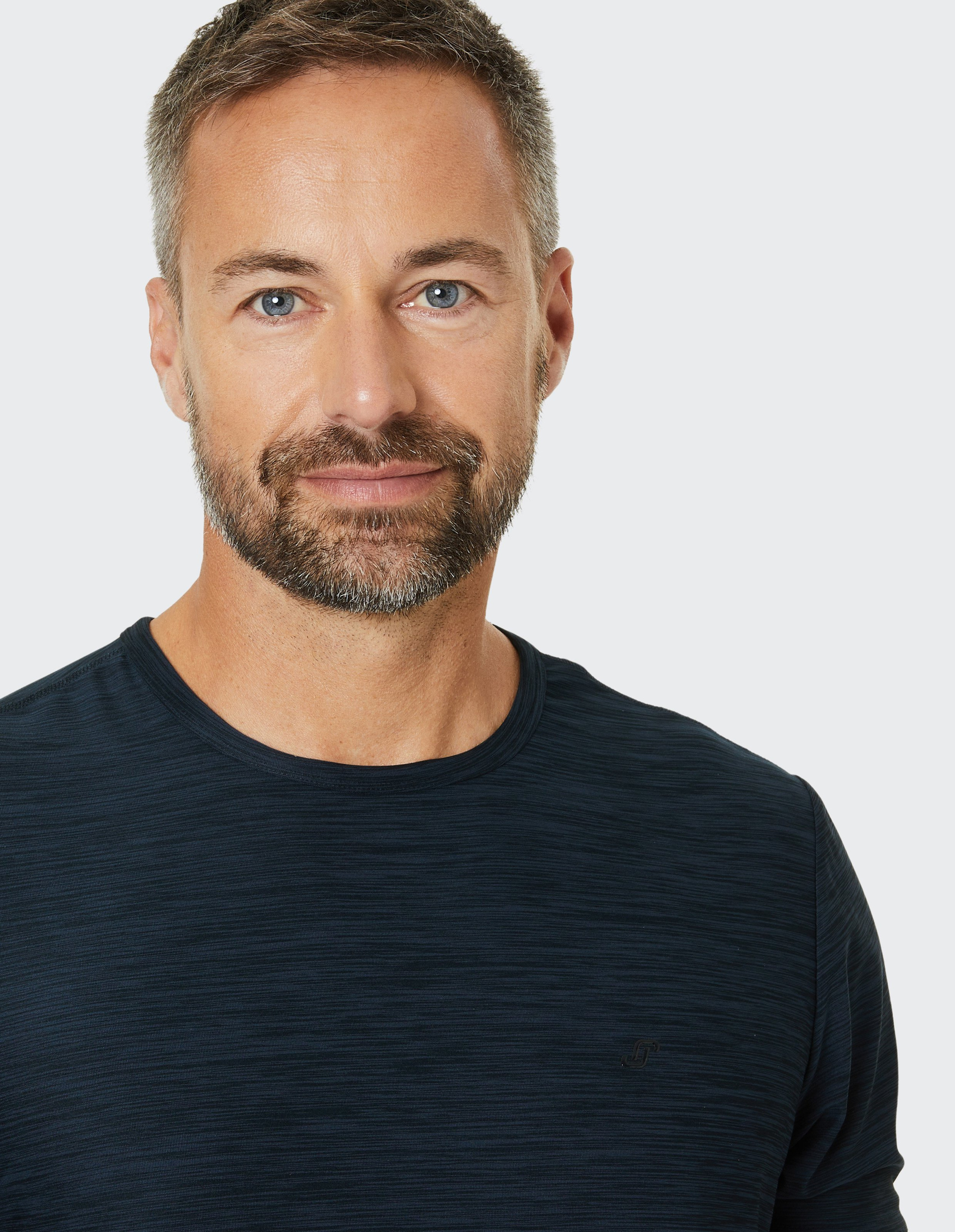 Ein lächelnder Mann mit Bart trägt ein dunkelblaues T-Shirt und schaut direkt in die Kamera, vor einem hellen Hintergrund.