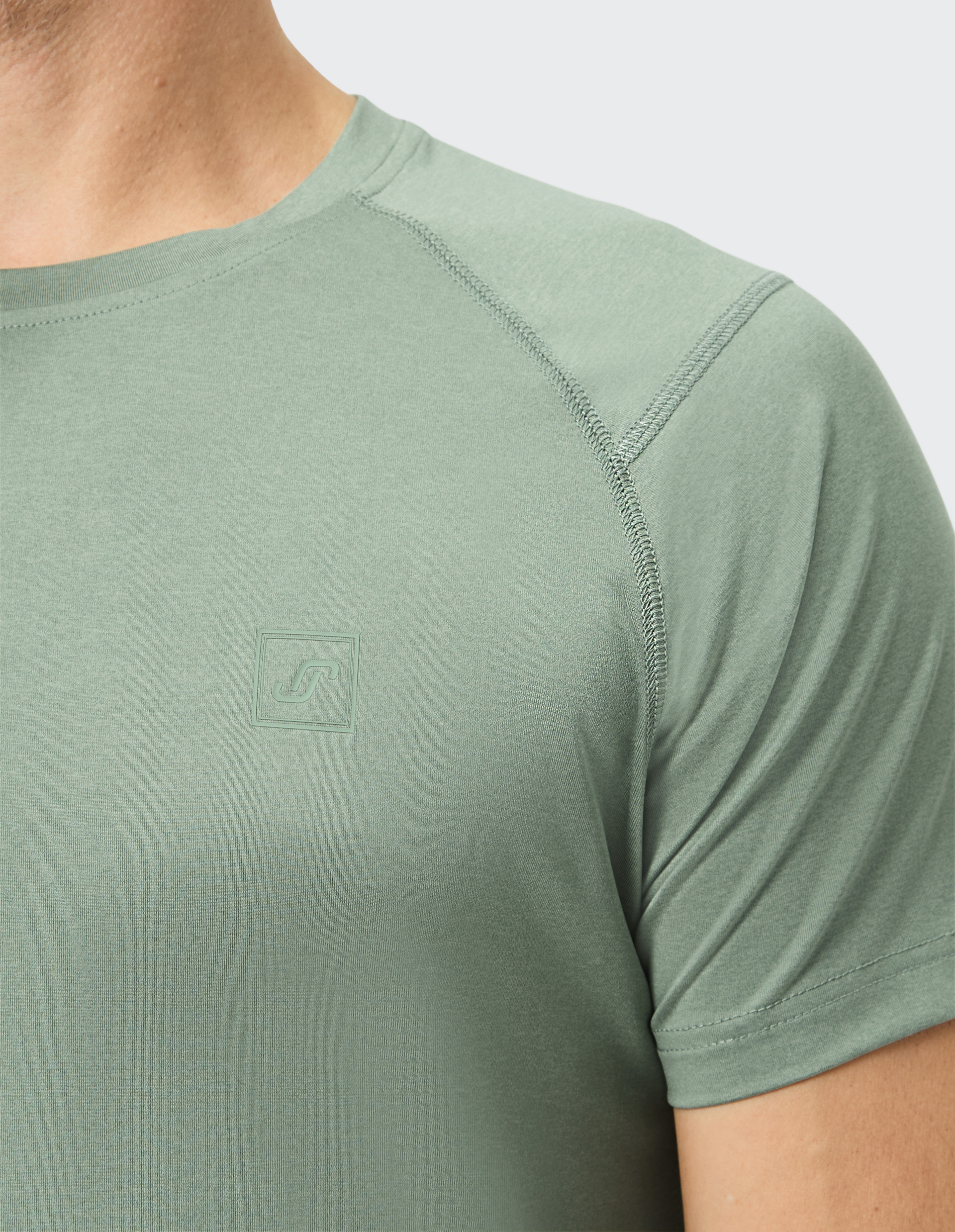 Grünes T-Shirt aus leichtem Stoff mit subtilem Logo auf der Brust, schrägen Nähten und kurzer Ärmelansicht, Schulter von Person sichtbar.
