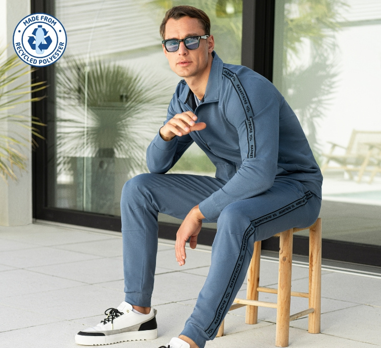 Mann in blaugrauem Anzug sitzt auf einem Hocker und macht eine sportliche Pose. In der Ecke des Bildes ist das Zeichen für Recycled Polyester angebracht.