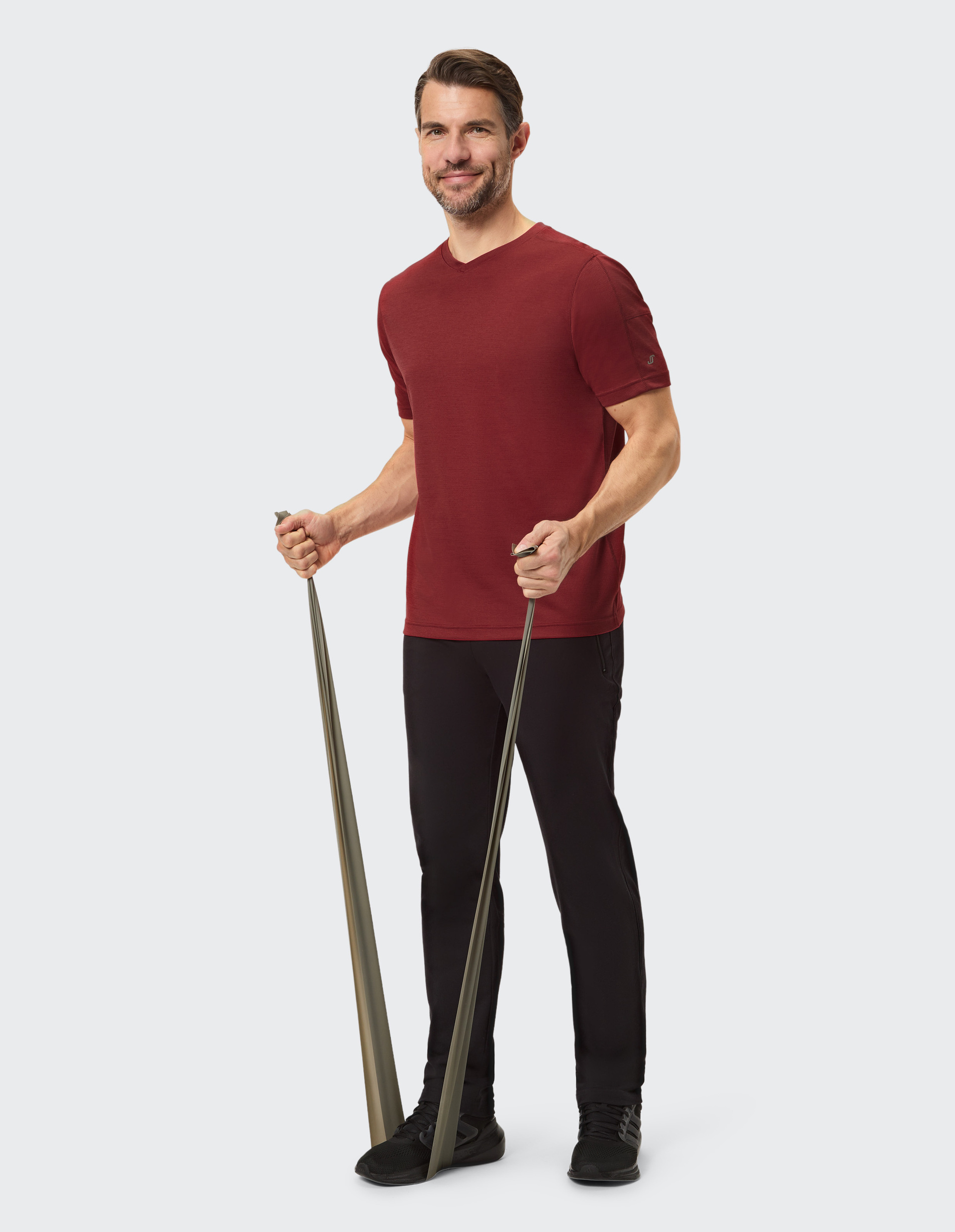 Ein Mann in dunkelrotem T-Shirt und schwarzen Jeans steht lächelnd, Hände locker an den Seiten, vor hellem Hintergrund.