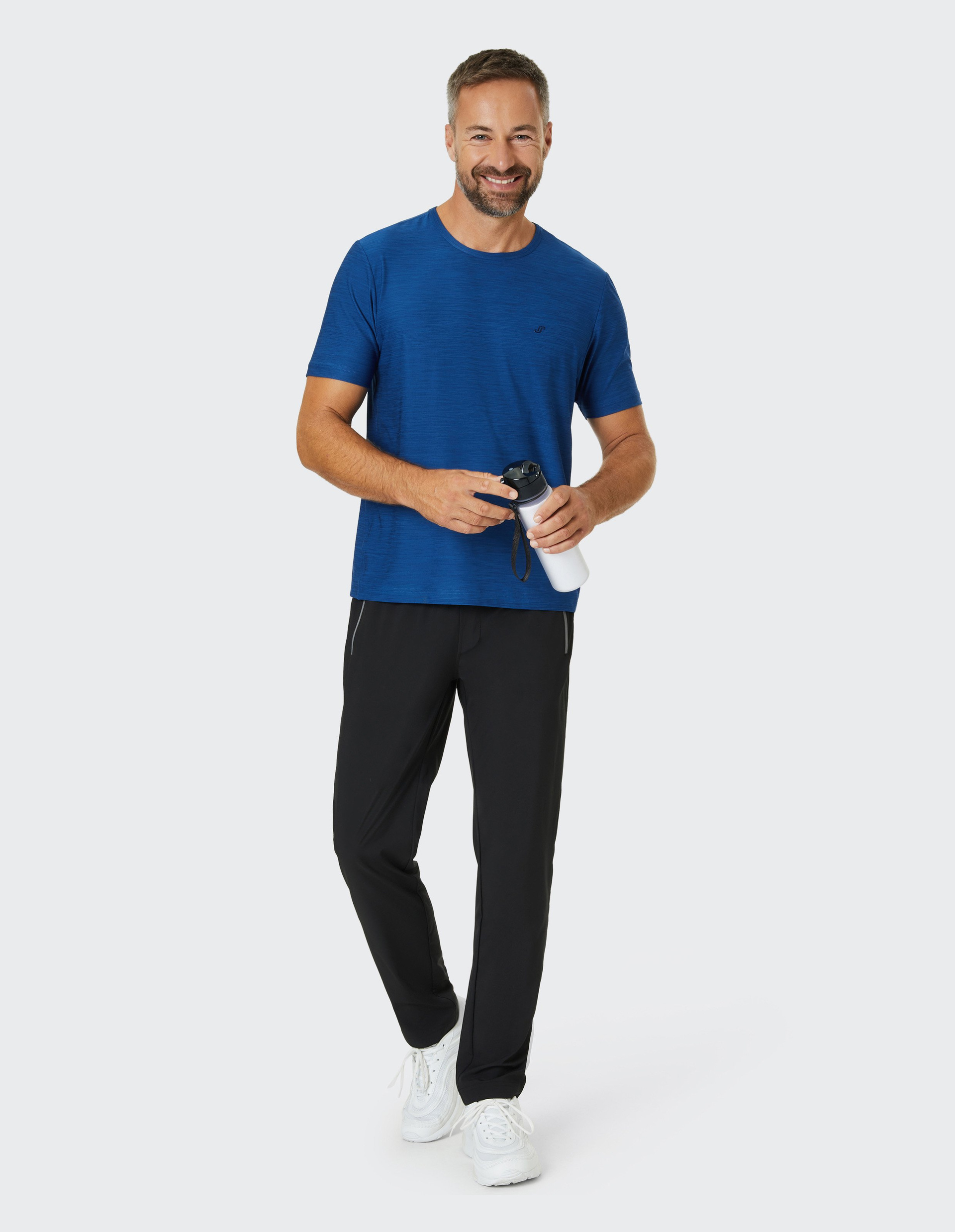 Ein Mann in blauem T-Shirt und schwarzer Hose hält eine Trinkflasche in der Hand, lächelnd vor hellem Hintergrund.