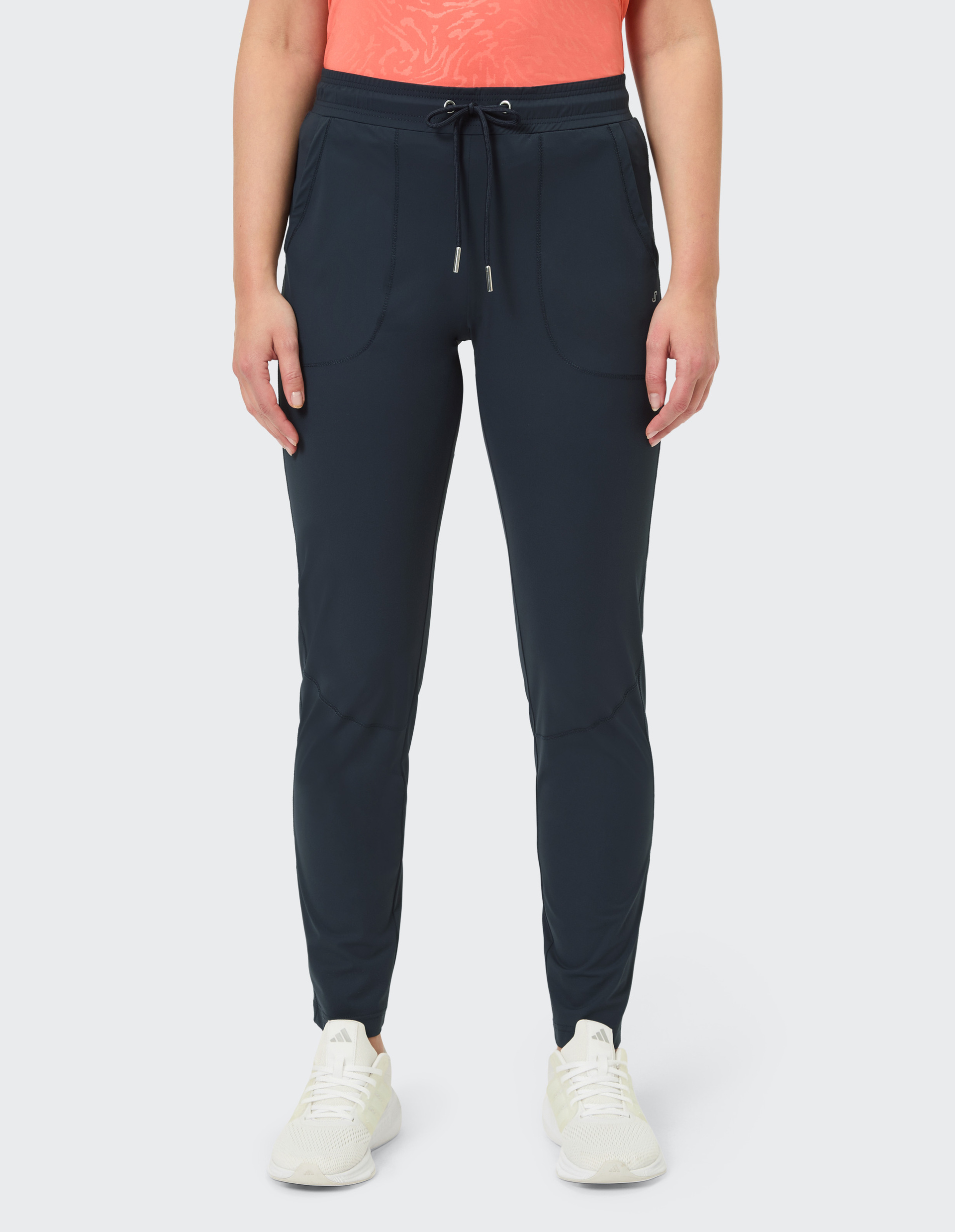 Person in dunkelblauer Jogginghose mit Kordelzug, orangefarbenem Oberteil und weißen Sportschuhen vor neutralem Hintergrund.