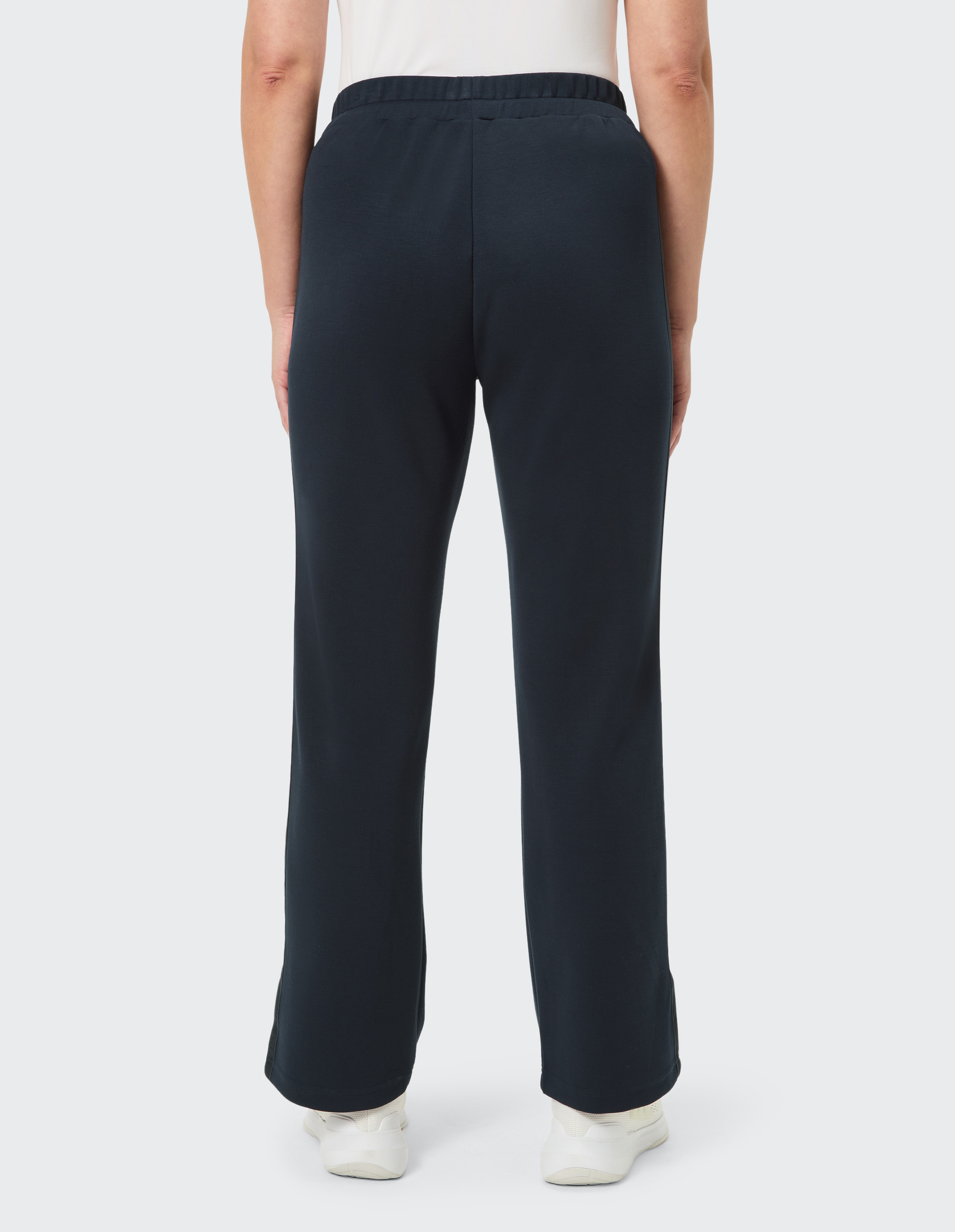 Eine Person trägt eine schwarze, locker sitzende Hose und weiße Turnschuhe. Die Ansicht zeigt die Rückseite der Beine.