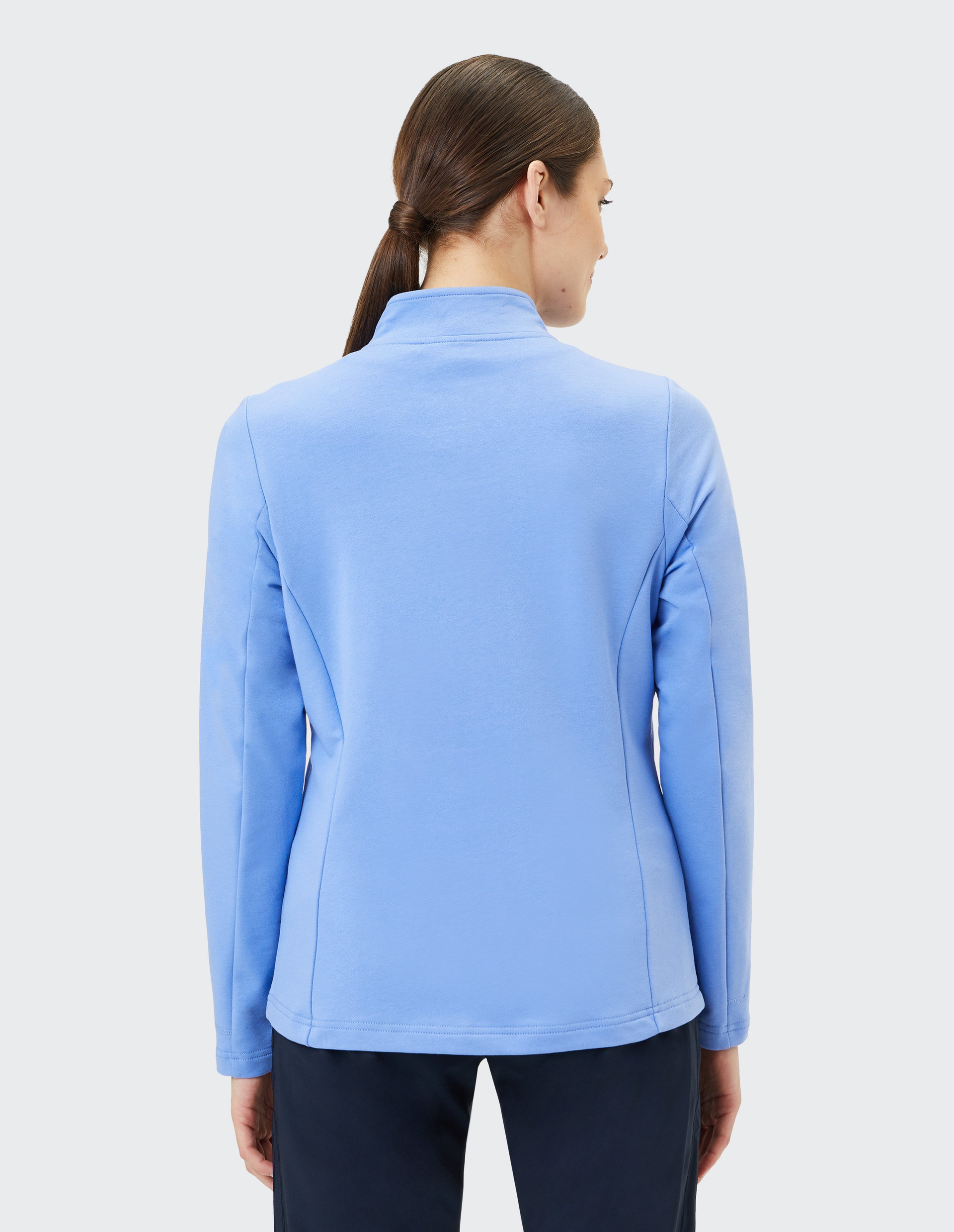 Frau mit braunem Haar im Zopf trägt hellblaue Jacke, steht mit dem Rücken zur Kamera vor neutralem Hintergrund.