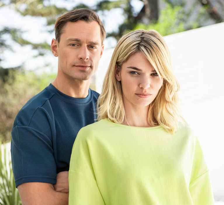 Mann in blauem Oberteil von JOY sportswear steht neben Frau in gelbem Oberteil von JOY sportswear. Beide sehen frontal in die Kamera.
