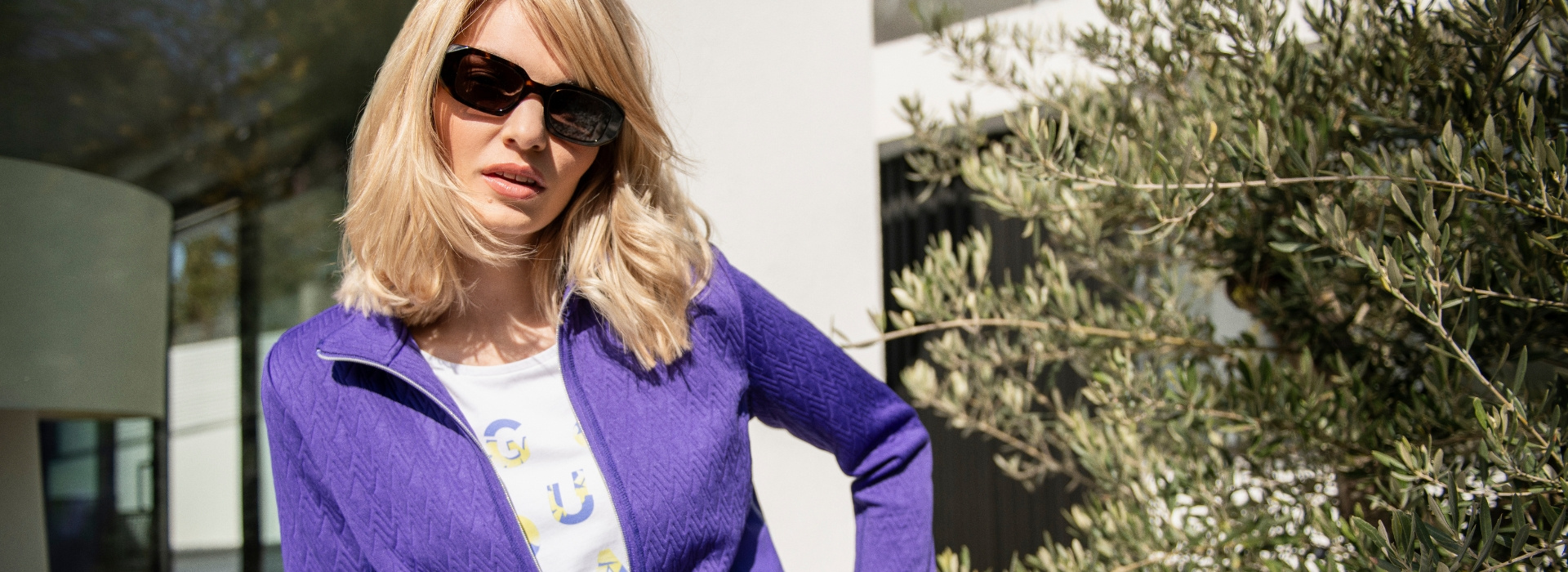 Frau in lilafarbener Jacke und Sonnenbrille bäugt sich zur Kamera.