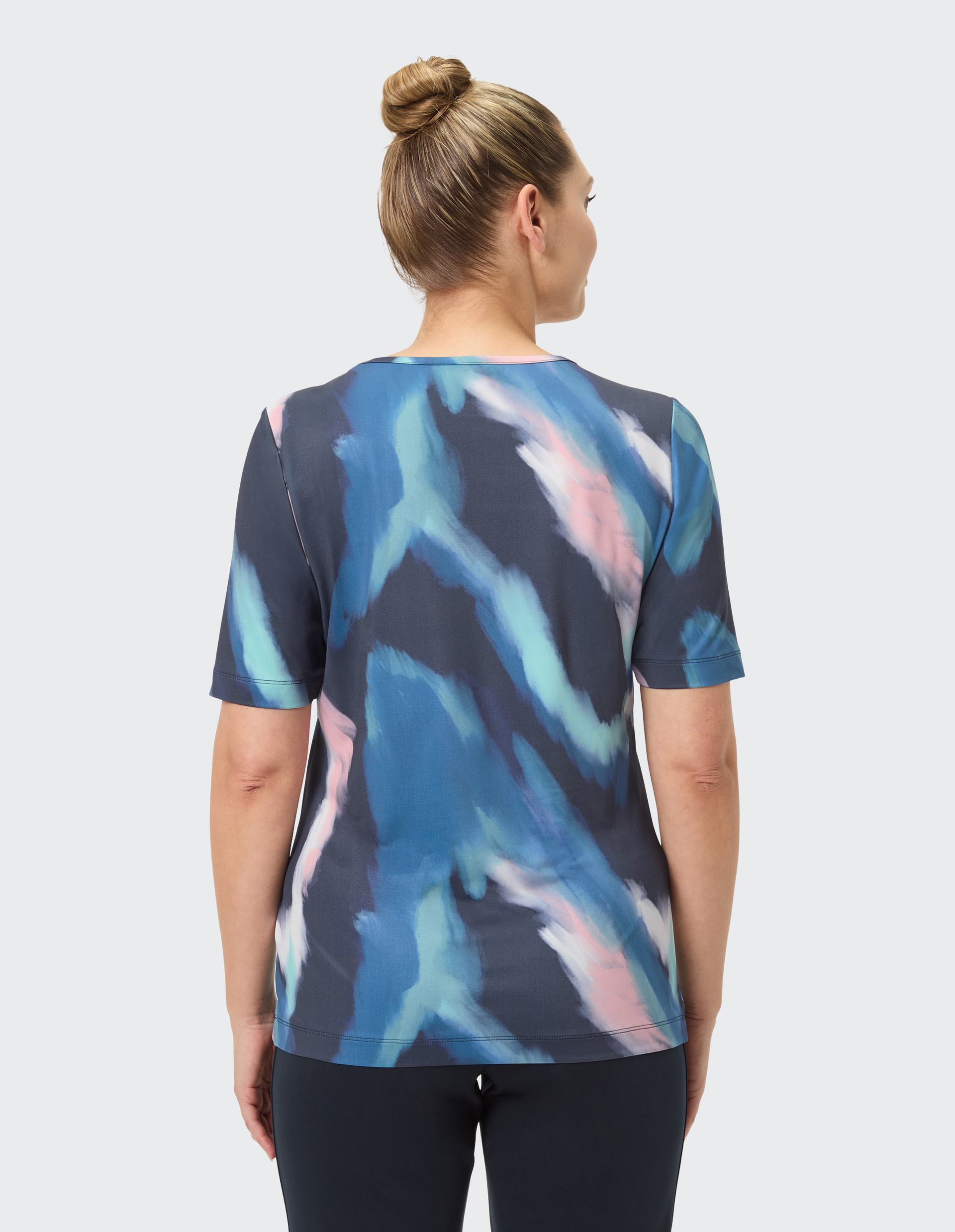 Frau von hinten, trägt ein T-Shirt mit blau-rosa Aquarellmuster und hat blondes, in einem Dutt hochgestecktes Haar.