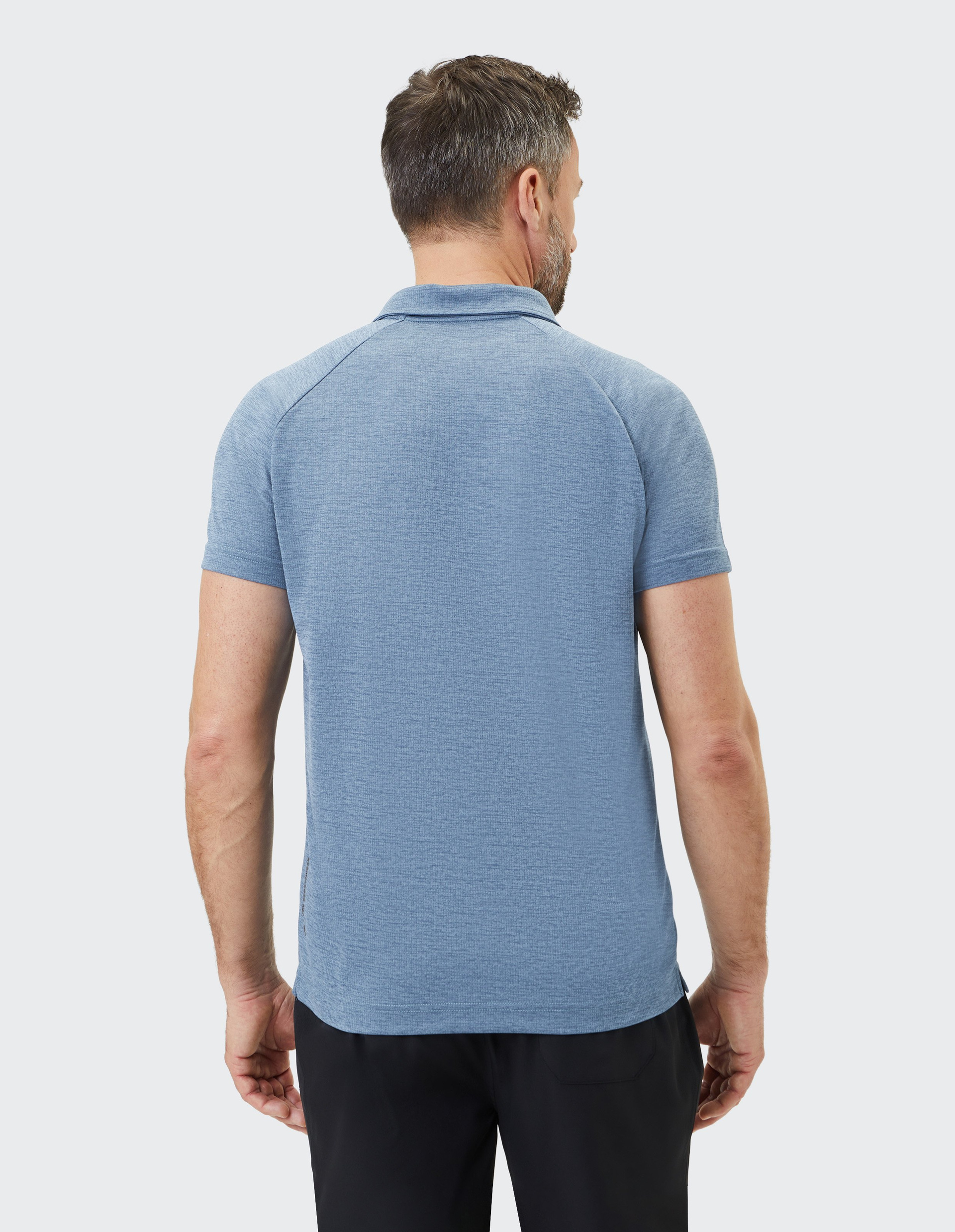 Ein Mann mit kurzen Haaren trägt ein blaues Poloshirt und schwarze Hosen, steht mit dem Rücken zur Kamera vor grauem Hintergrund.