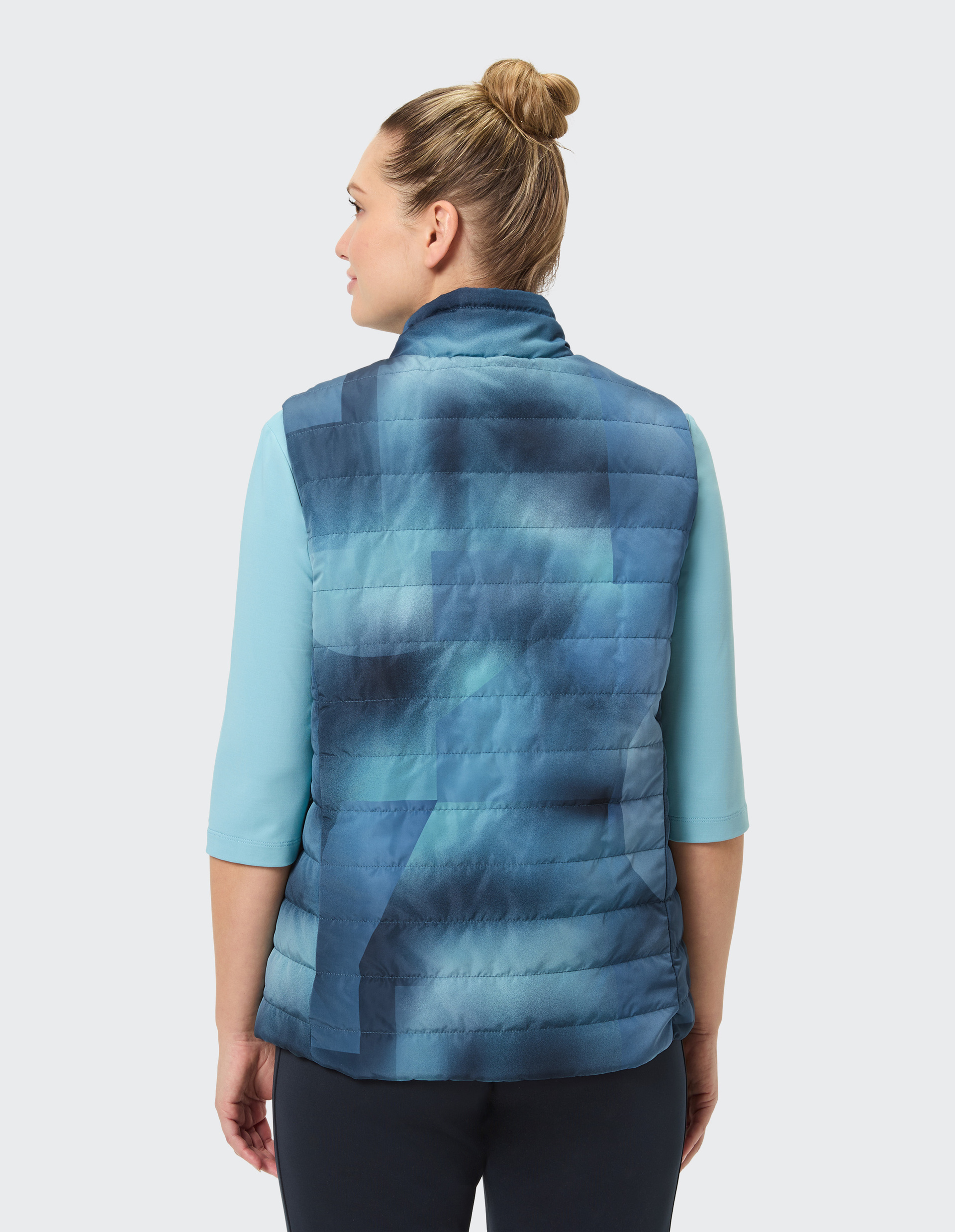 Eine Person mit hochgestecktem Haar trägt eine blau gemusterte Weste und ein hellblaues Shirt, von hinten fotografiert.