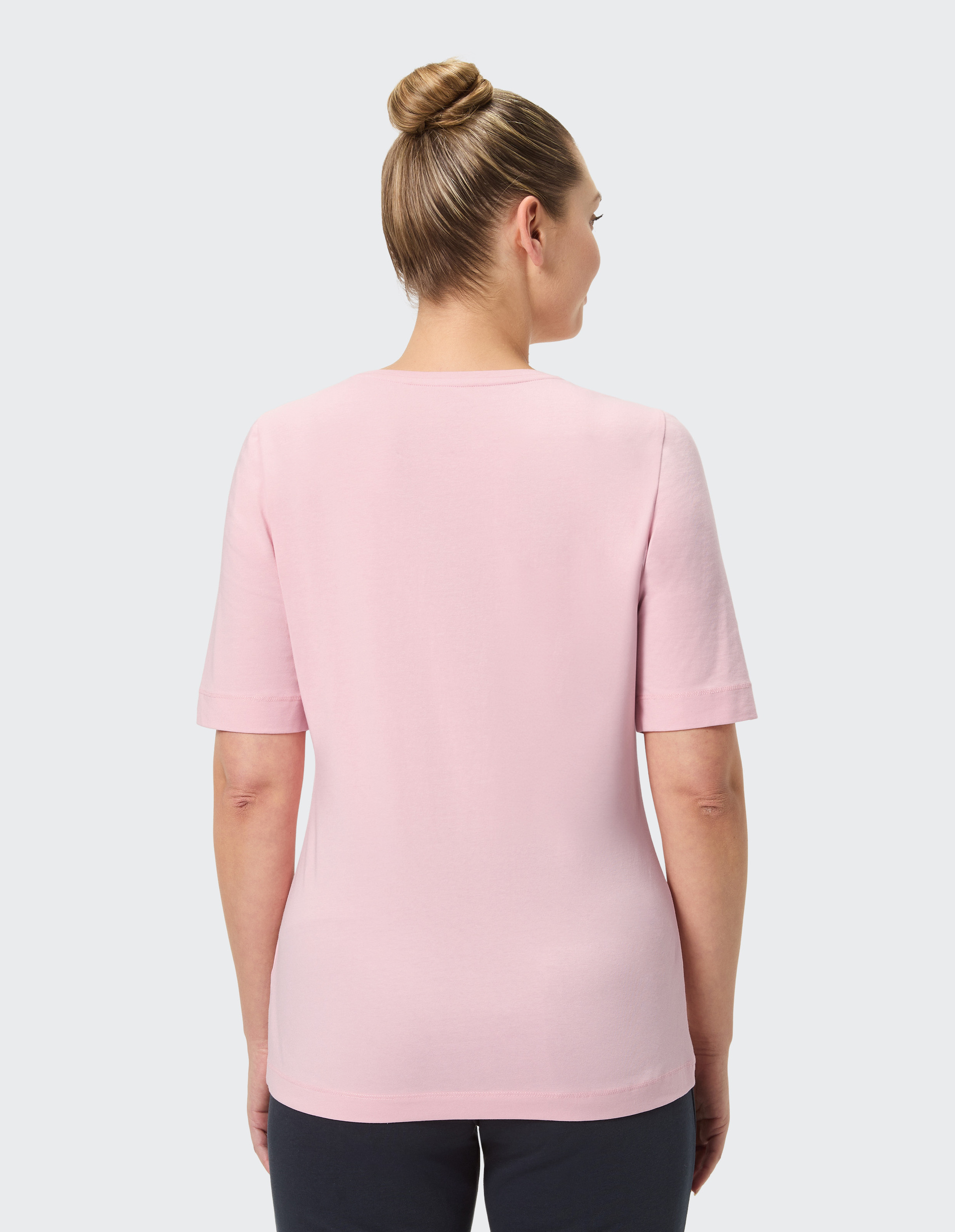 Eine Person von hinten in einem rosa T-Shirt, die Haare zu einem Dutt gebunden, trägt dunkelblaue Hosen, vor grauem Hintergrund.